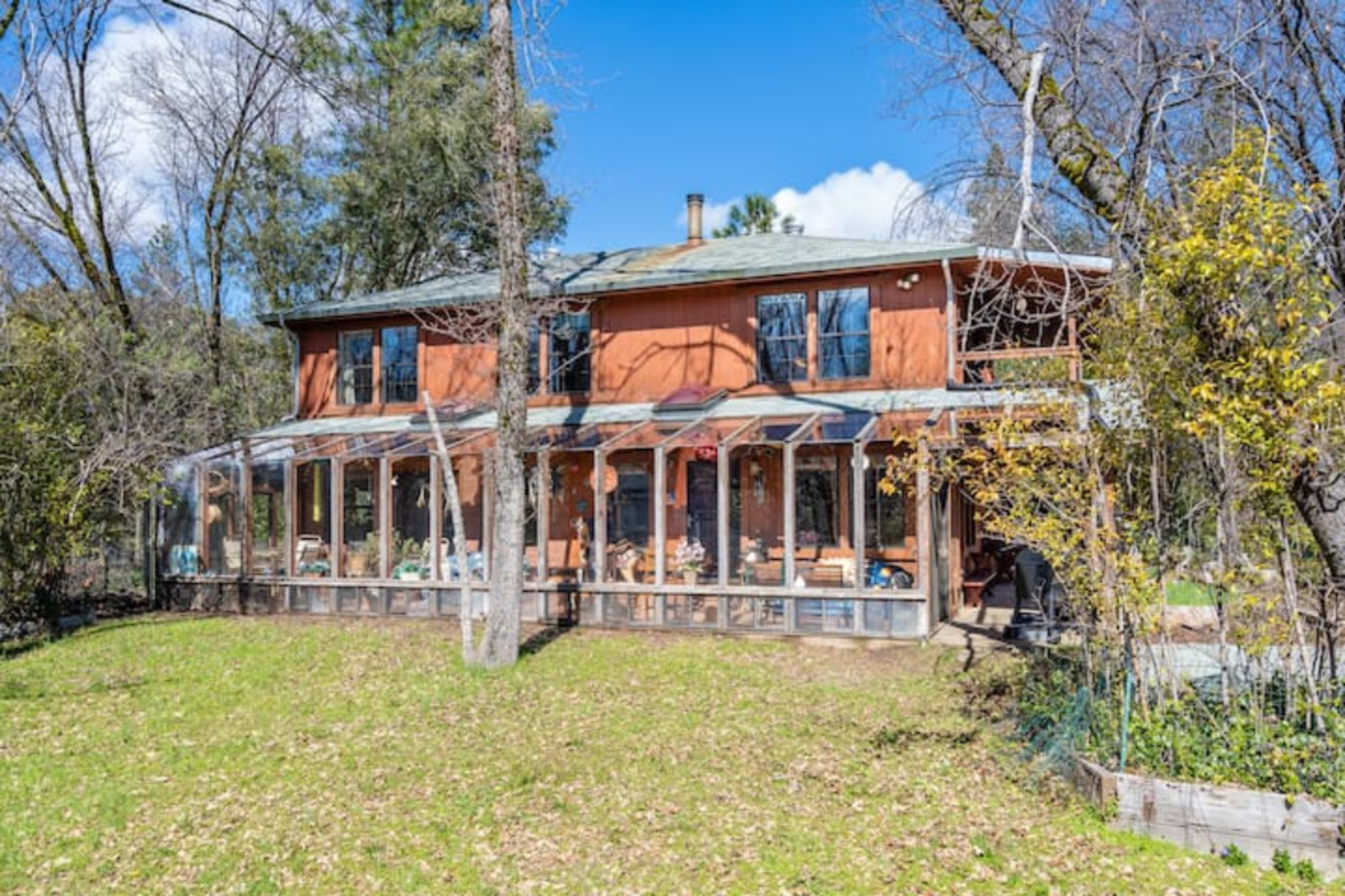 A two-story house with a reddish-orange exterior features a large glass-enclosed porch and is surrounded by trees and greenery.