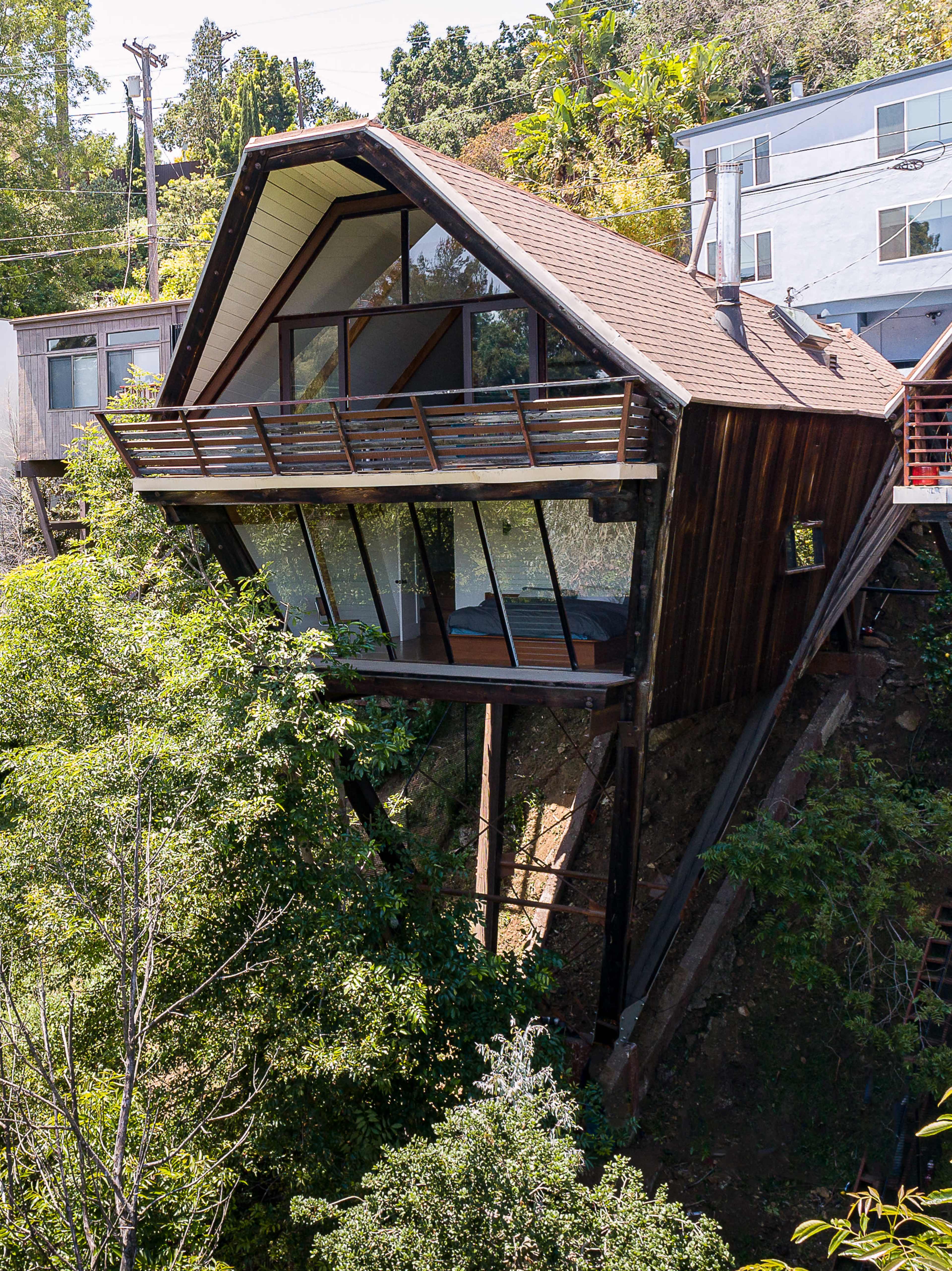 A modern, angular house with large glass windows is perched on stilts above a hillside, surrounded by lush greenery.