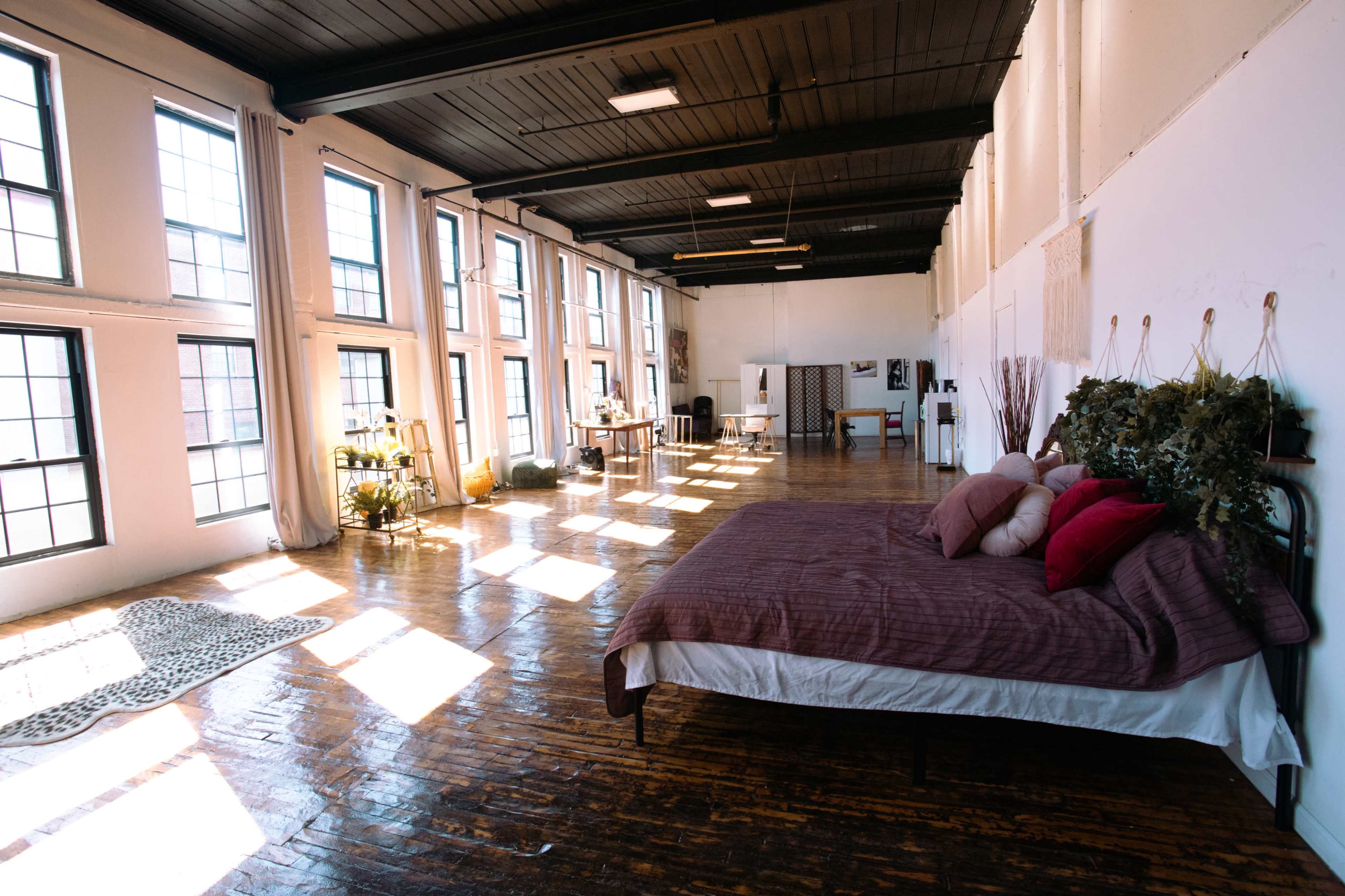 A spacious loft with large windows features a bed adorned with decorative pillows and a wooden floor reflecting sunlight.
