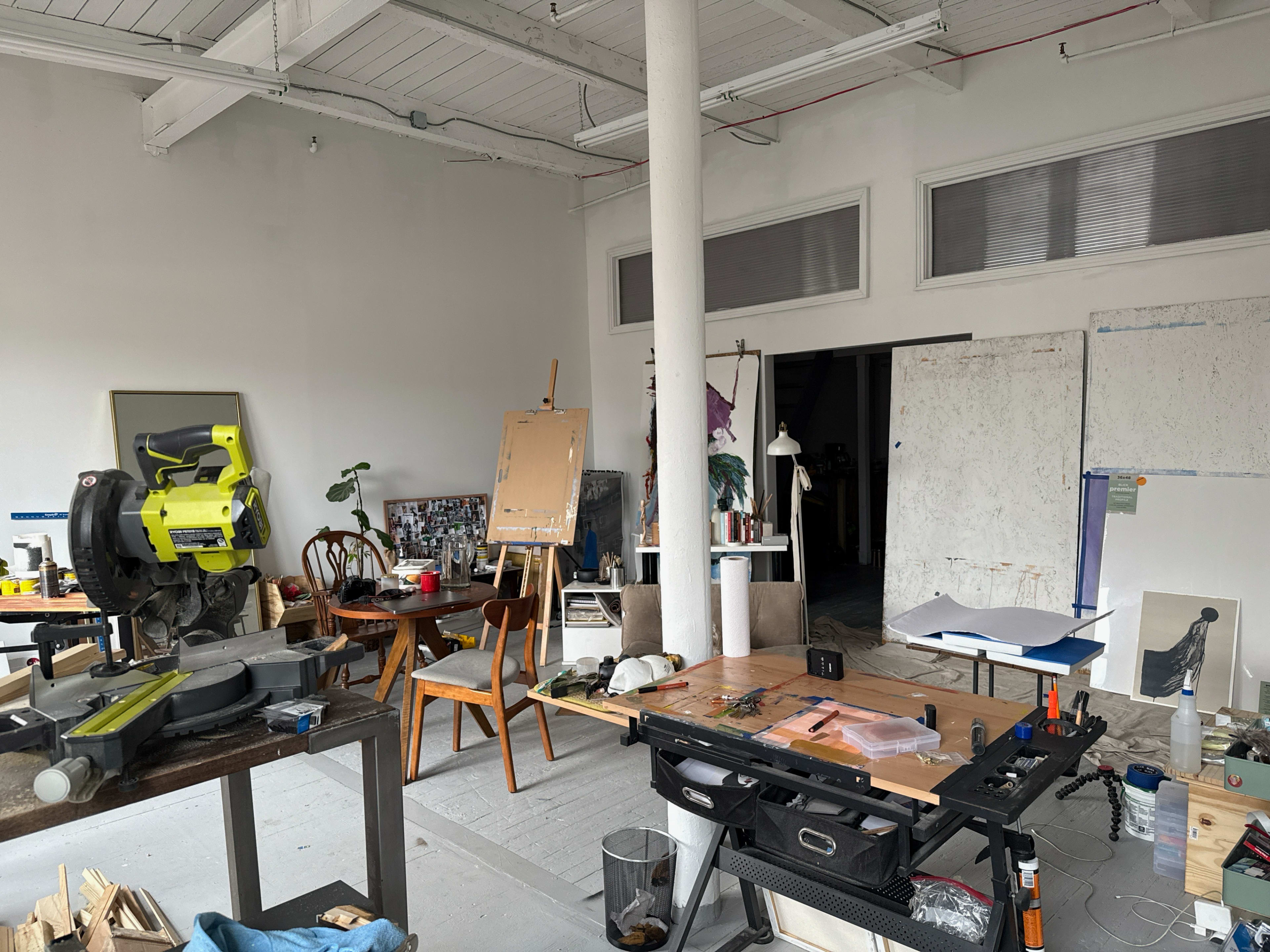 The image shows a cluttered workshop with various tools, furniture, and art supplies scattered throughout the space.