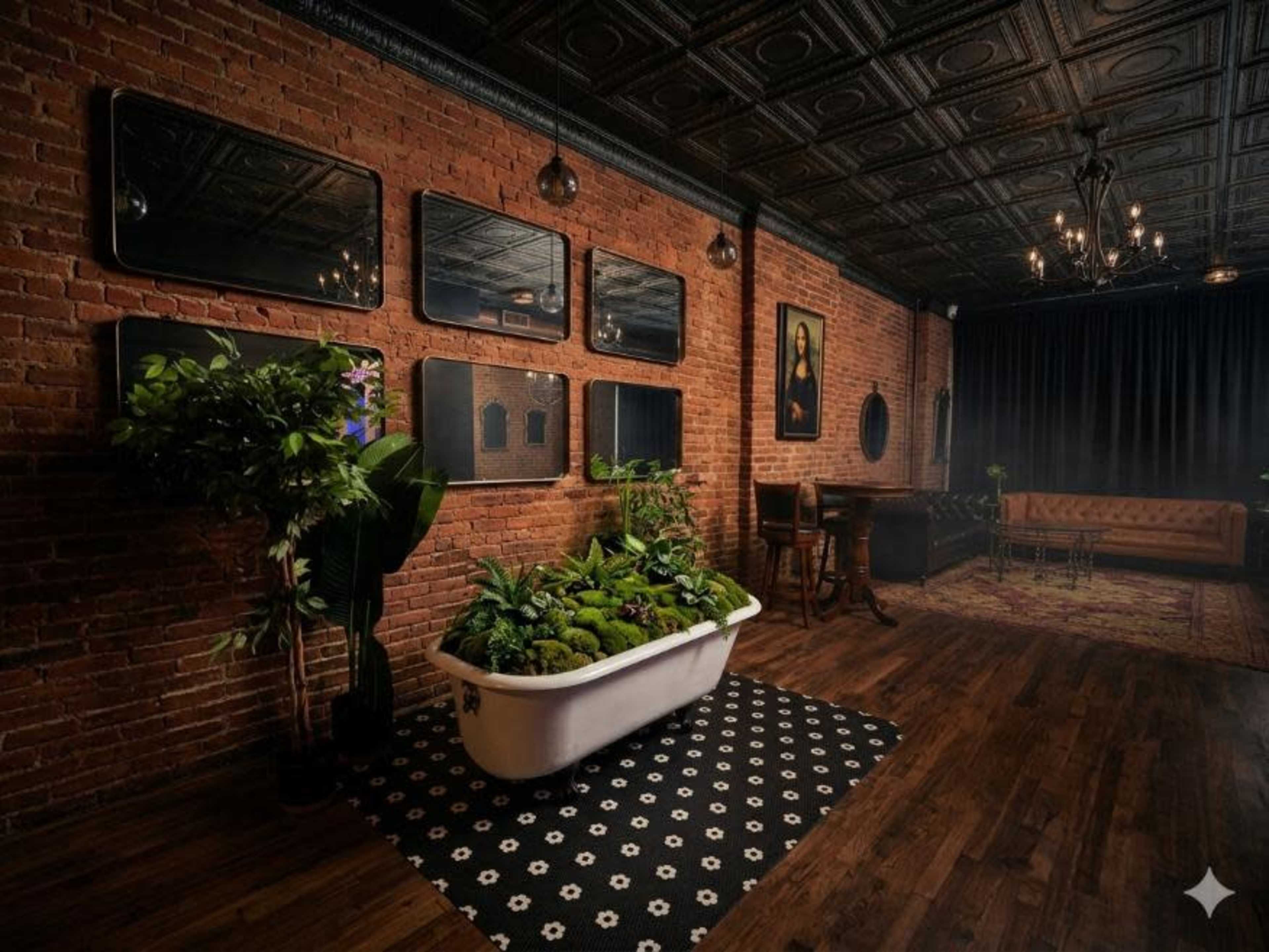 The image shows a room with exposed brick walls, dark wood flooring, a bathtub filled with plants, framed mirrors, and a vintage-style sofa.