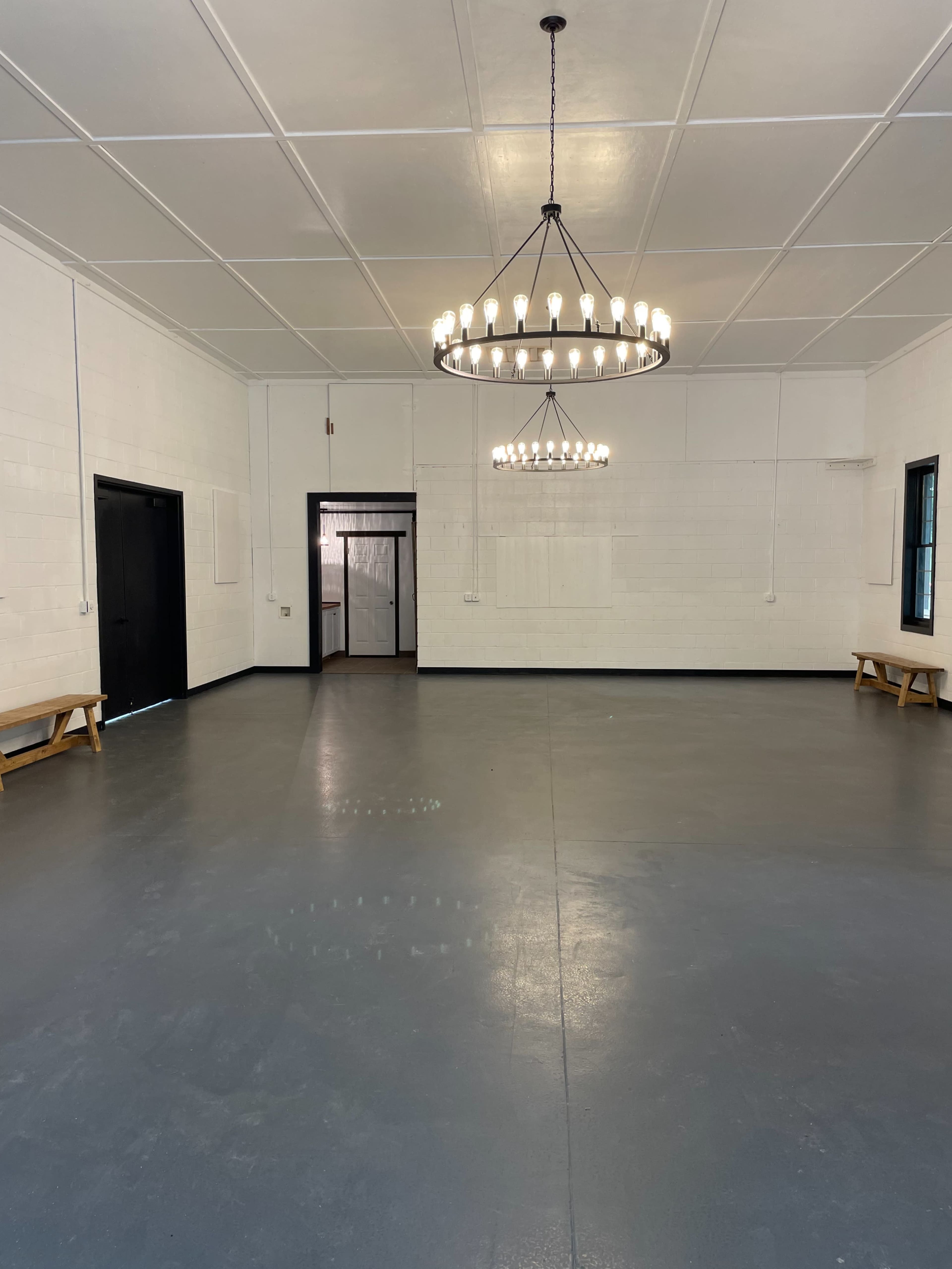 A spacious room with a smooth gray floor, two large chandeliers hanging from the ceiling, and simple wooden benches along the walls.