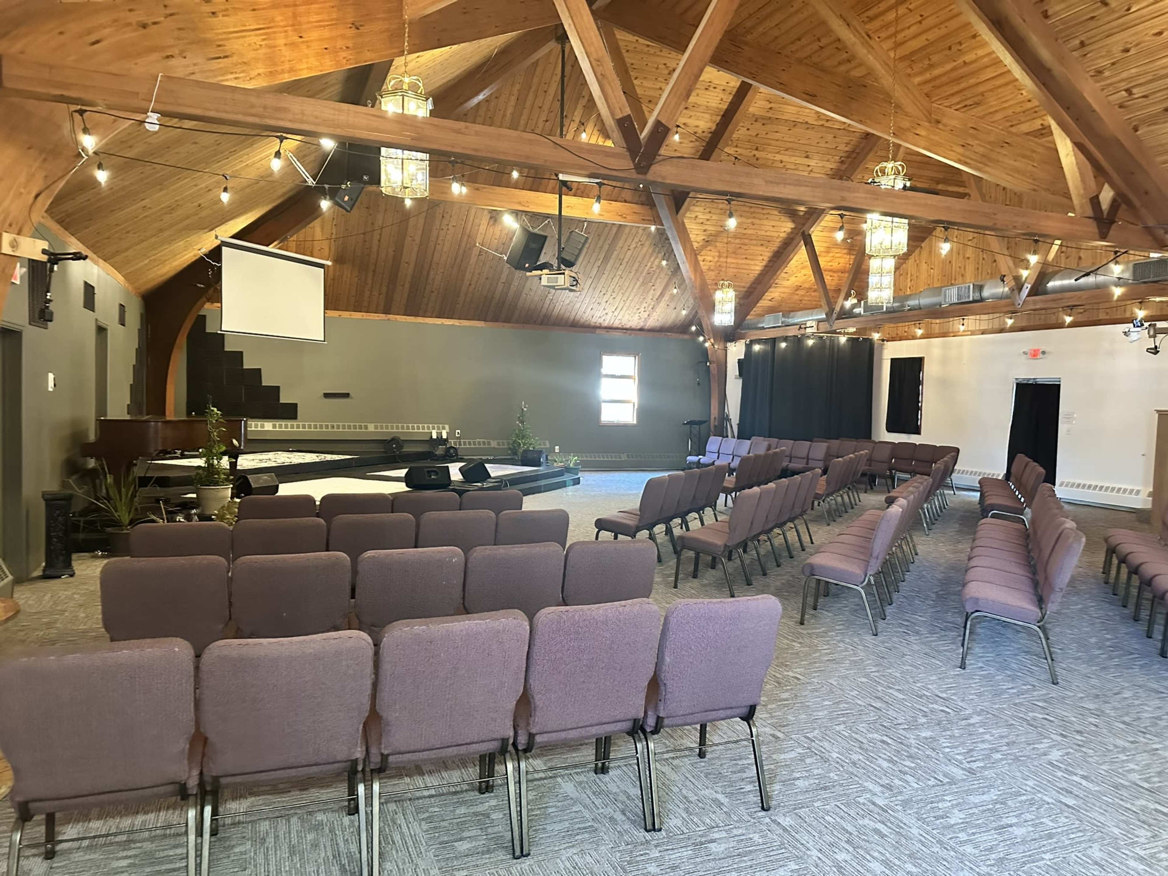 An interior of a spacious auditorium with wooden beams, rows of chairs facing a stage, and a large projection screen.