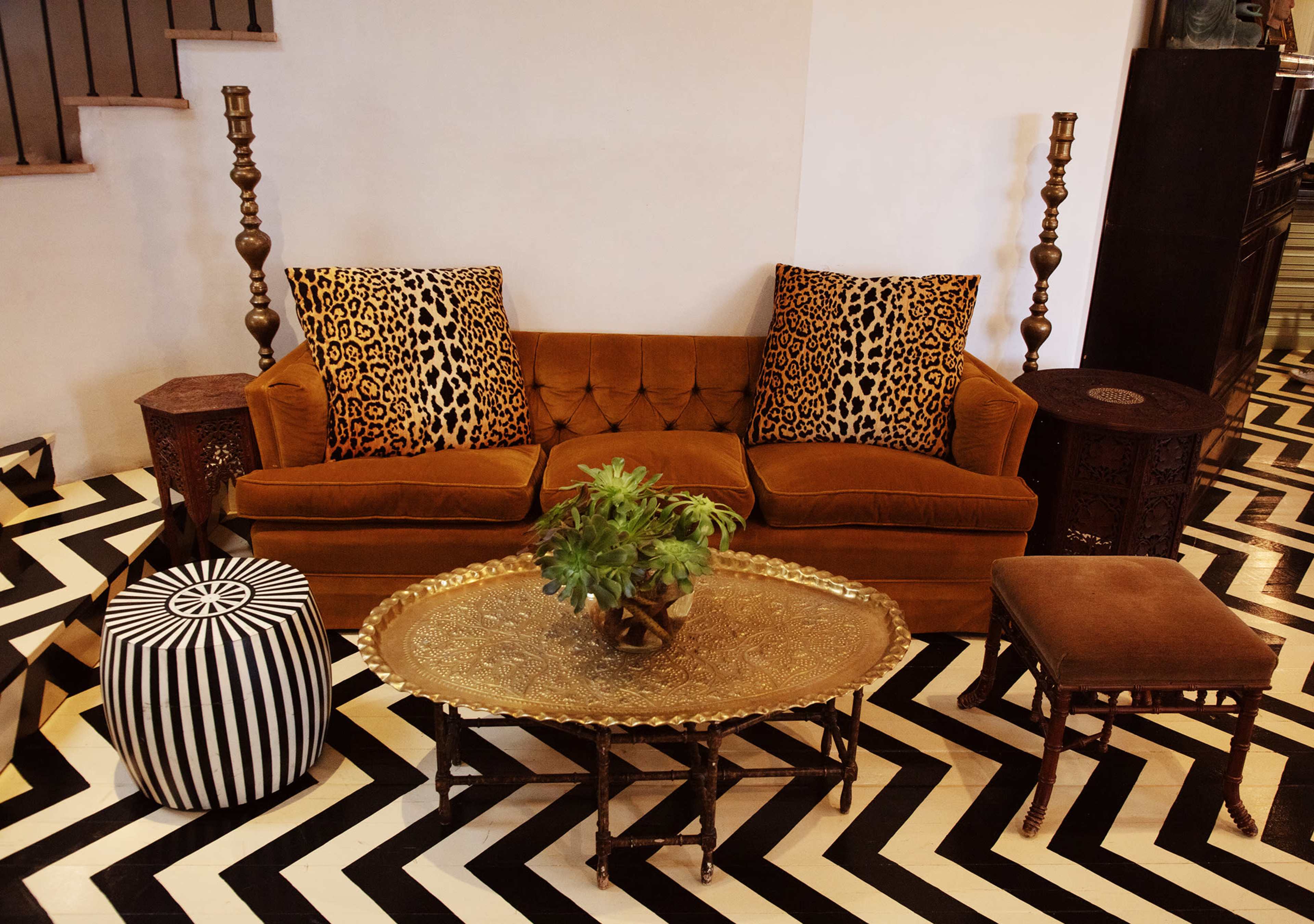 The image shows a stylish living room with a brown velvet sofa, decorative pillows, a round coffee table with intricate design, and contrasting black and white zigzag flooring.