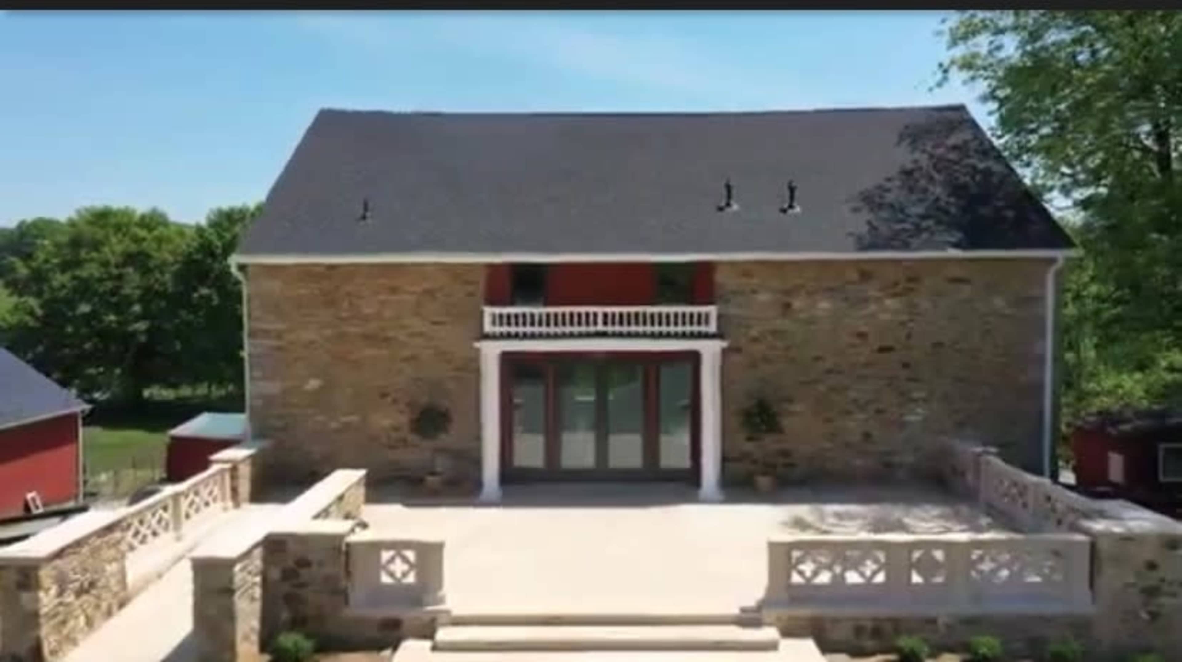 A large, stone building with a black roof features a spacious terrace and double doors at the front, surrounded by greenery.