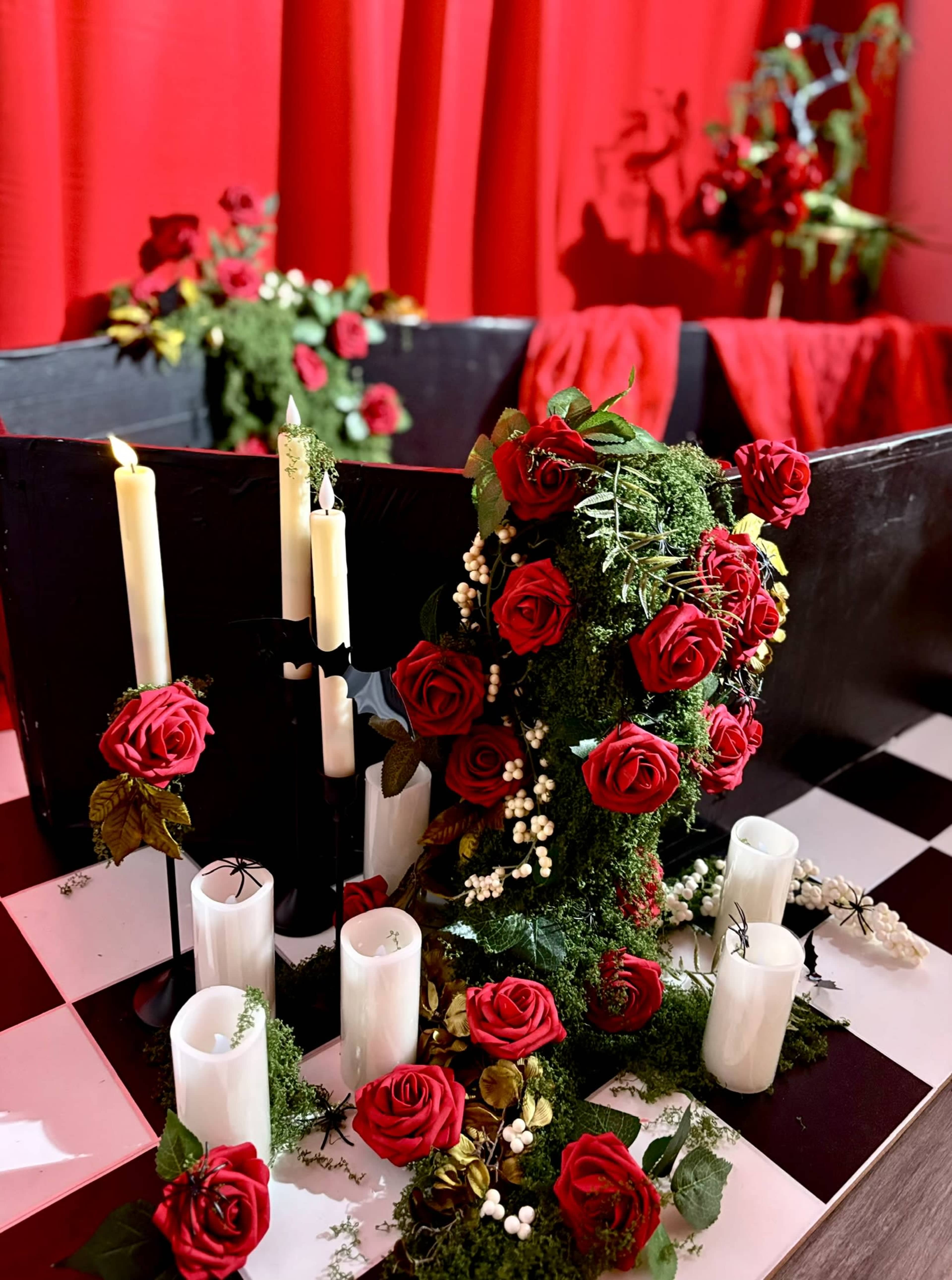 A decorative arrangement featuring red roses, white candles, and greenery set against a red backdrop.
