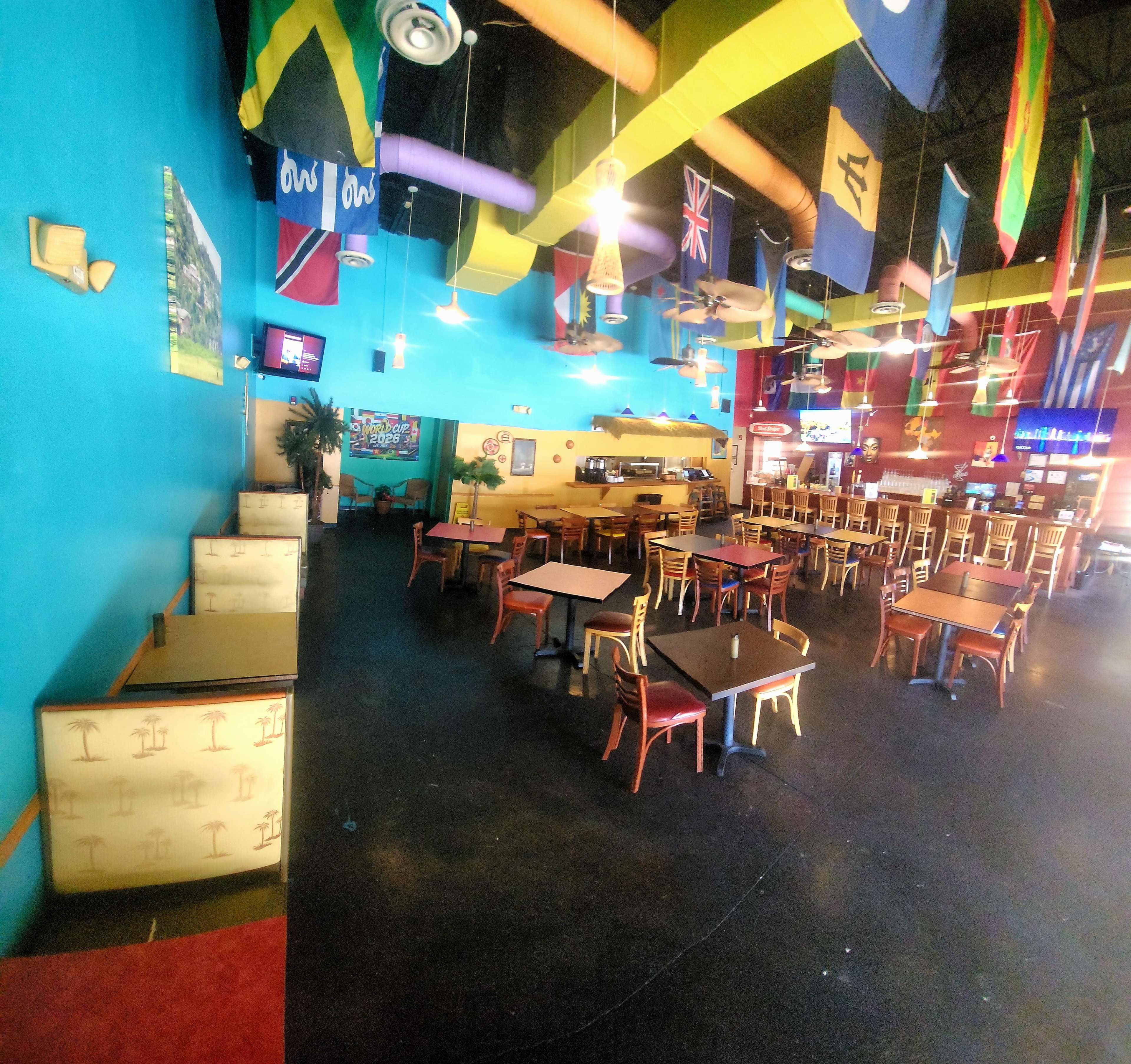 The image shows a brightly colored restaurant interior with empty tables and chairs, adorned with various international flags hanging from the ceiling.