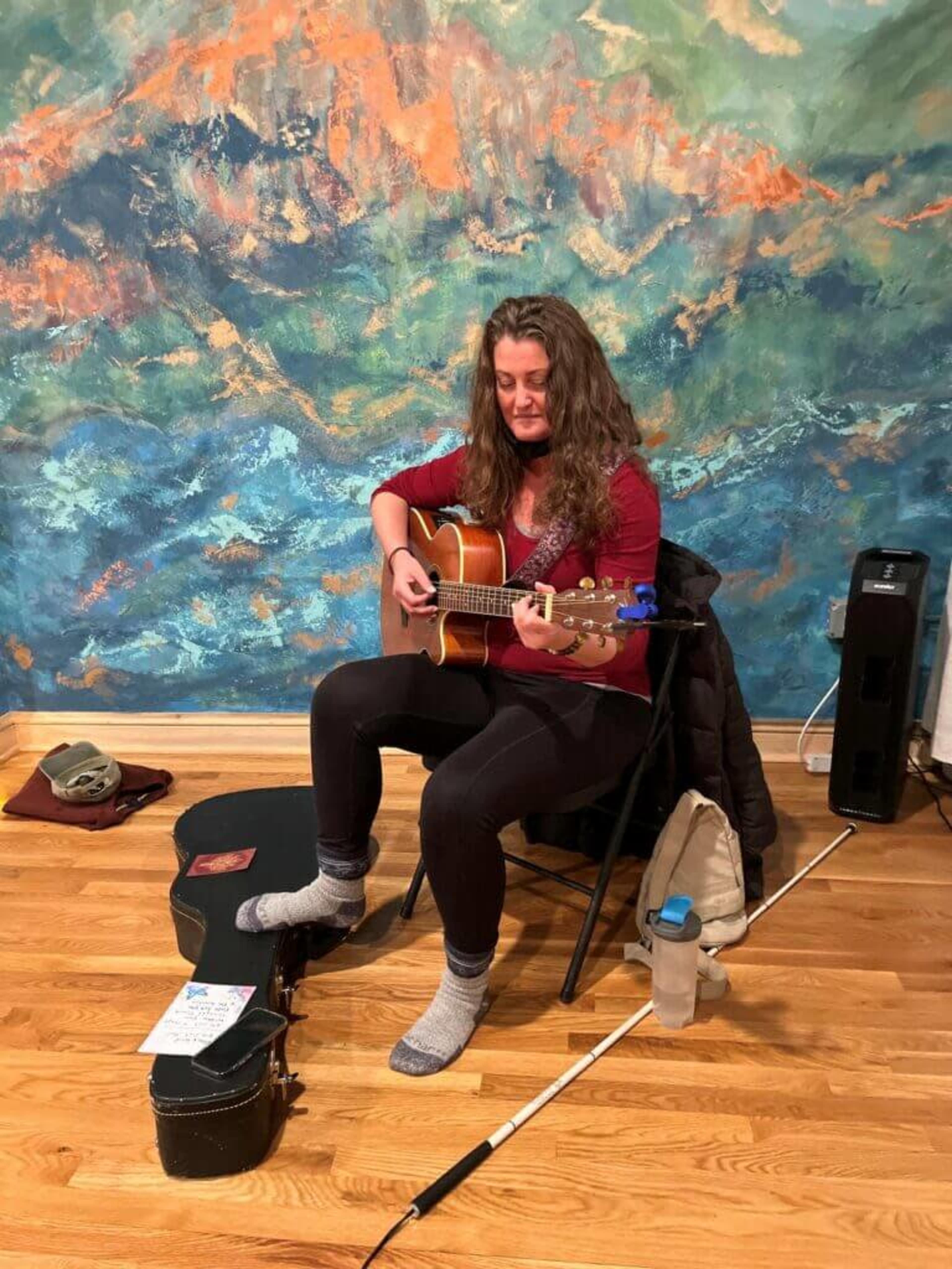 A woman sits on a folding chair playing an acoustic guitar in front of a colorful mural.