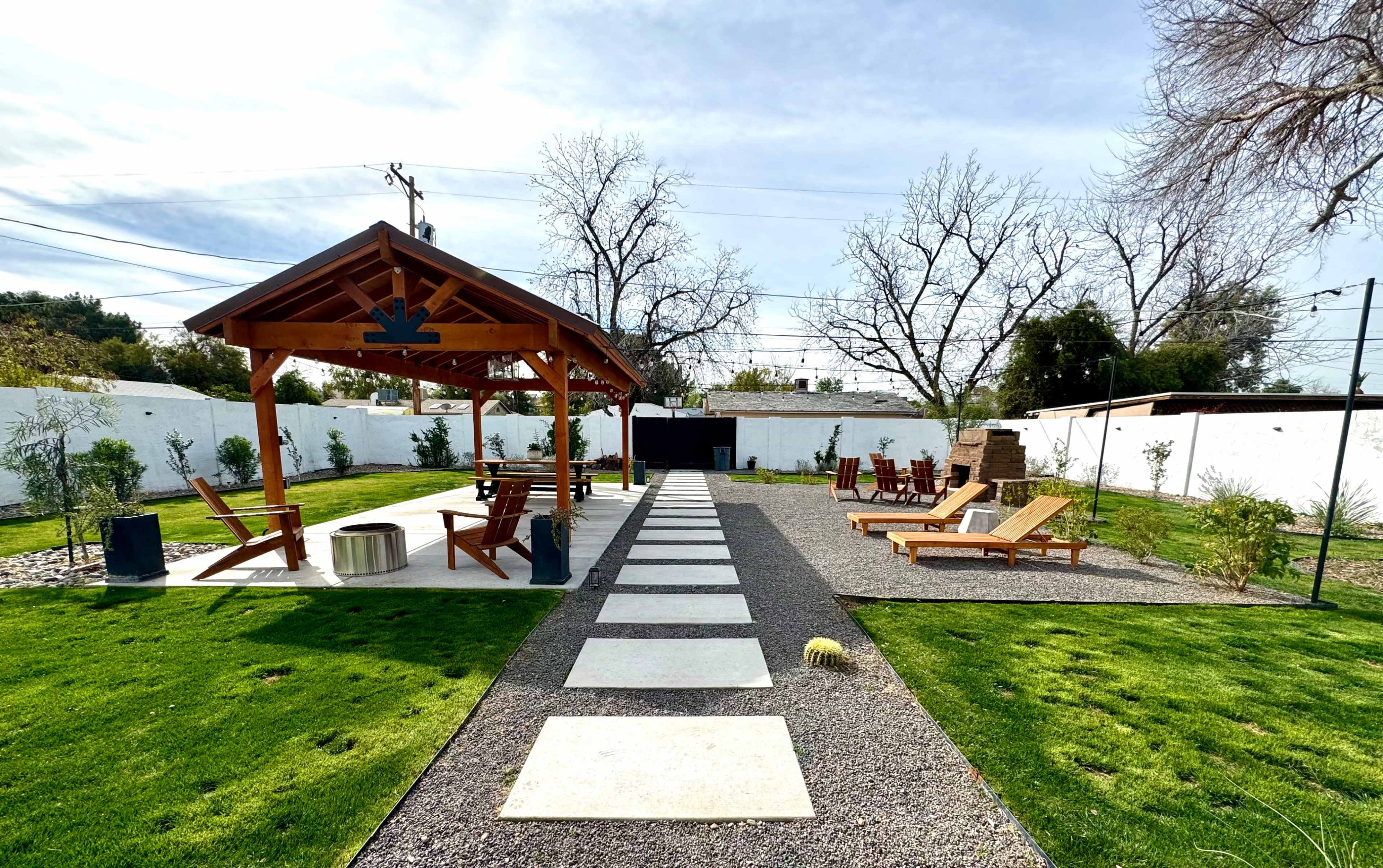 A landscaped outdoor space with a wooden pavilion, a stone pathway, lounging chairs, and greenery along a white wall.