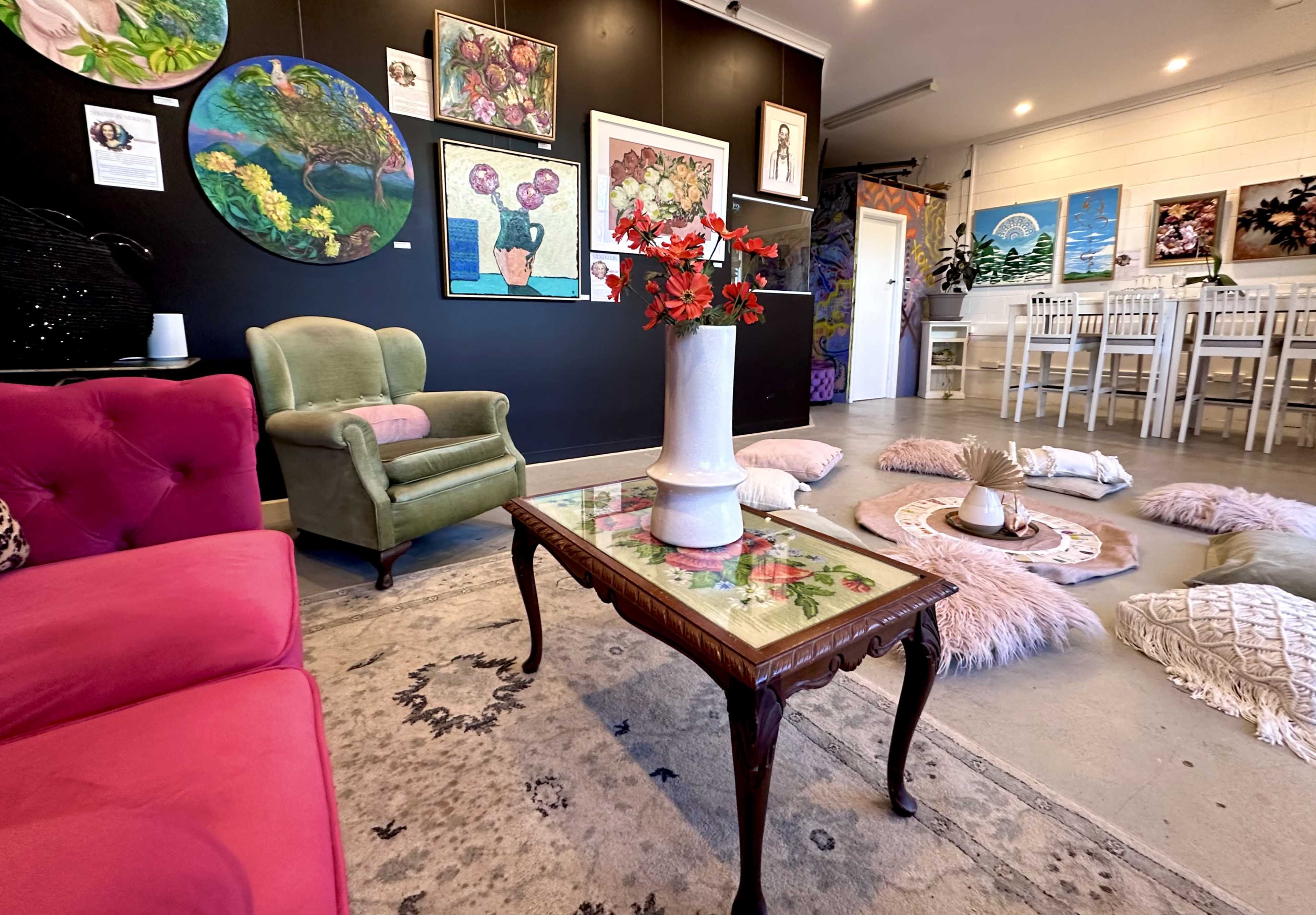 A cozy gallery space featuring colorful artwork on dark walls, a pink sofa, a green armchair, and a decorative table in the center surrounded by soft cushions.