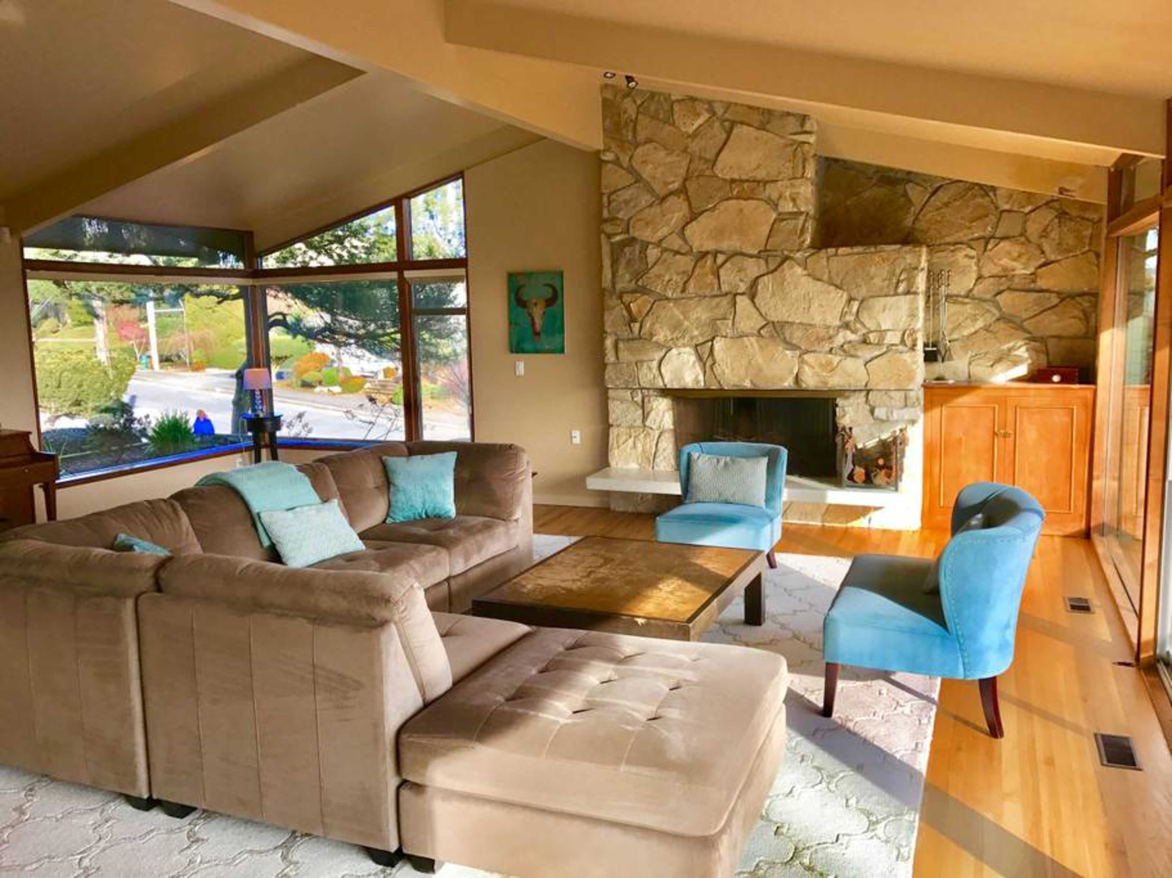 A cozy living room features a stone fireplace, a sectional sofa, and two blue accent chairs arranged around a wooden coffee table.