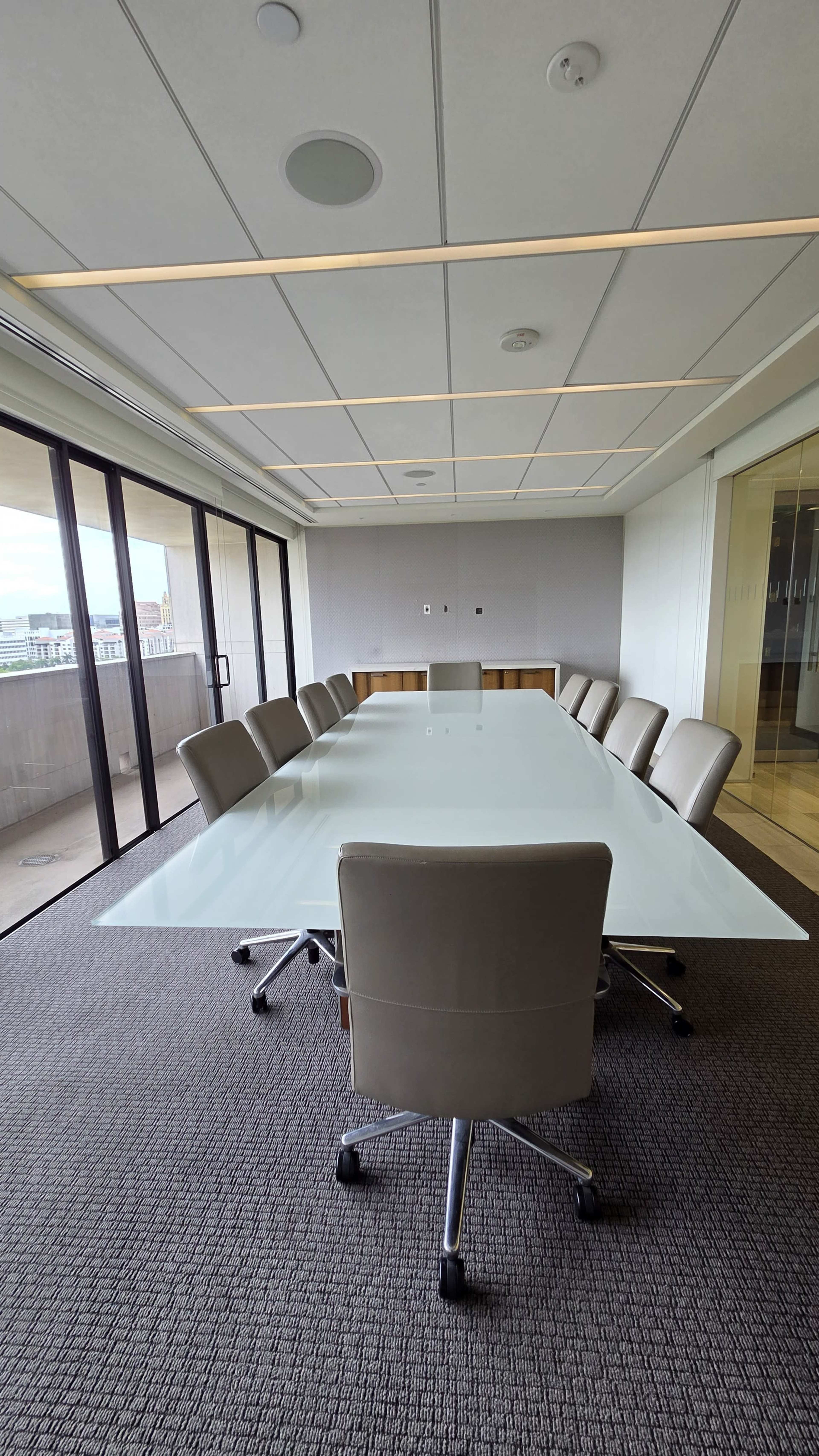 A modern conference room features a large glass table surrounded by eight leather chairs, with walls of windows allowing natural light.