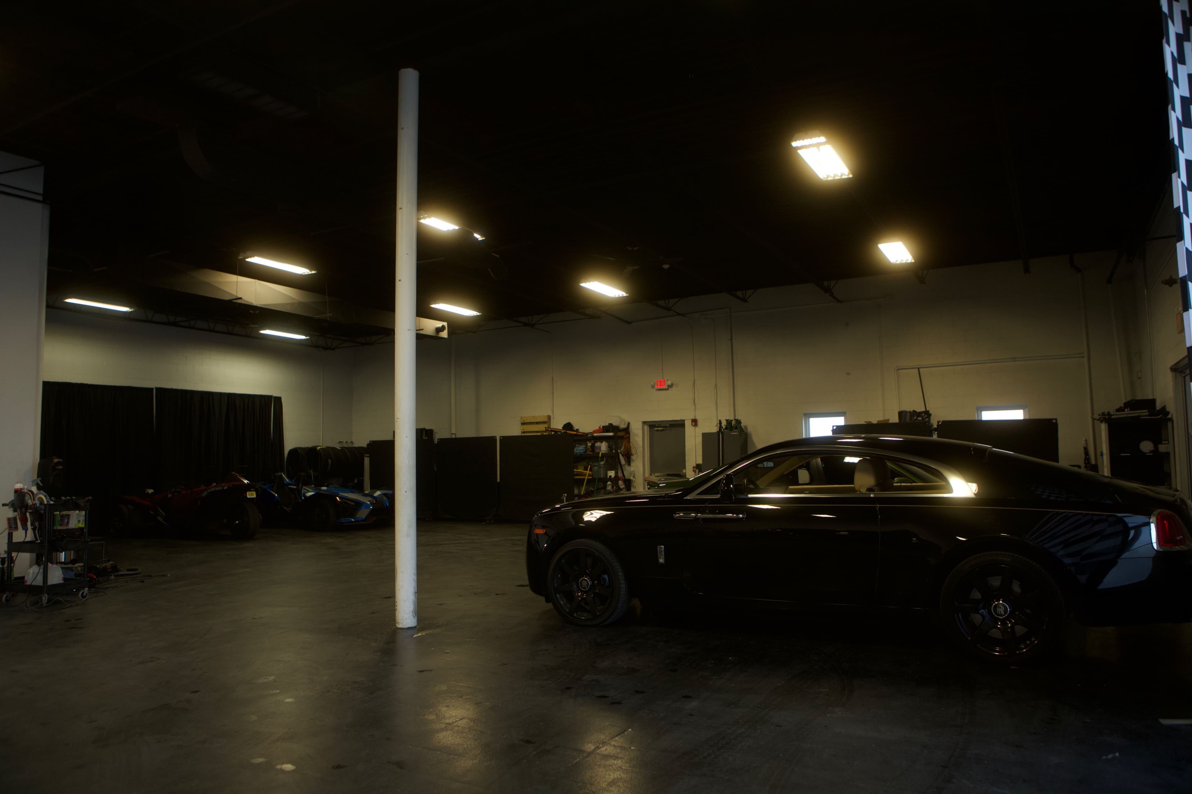 A black car is parked on the right side of a dimly lit garage with various tools and vehicles in the background.