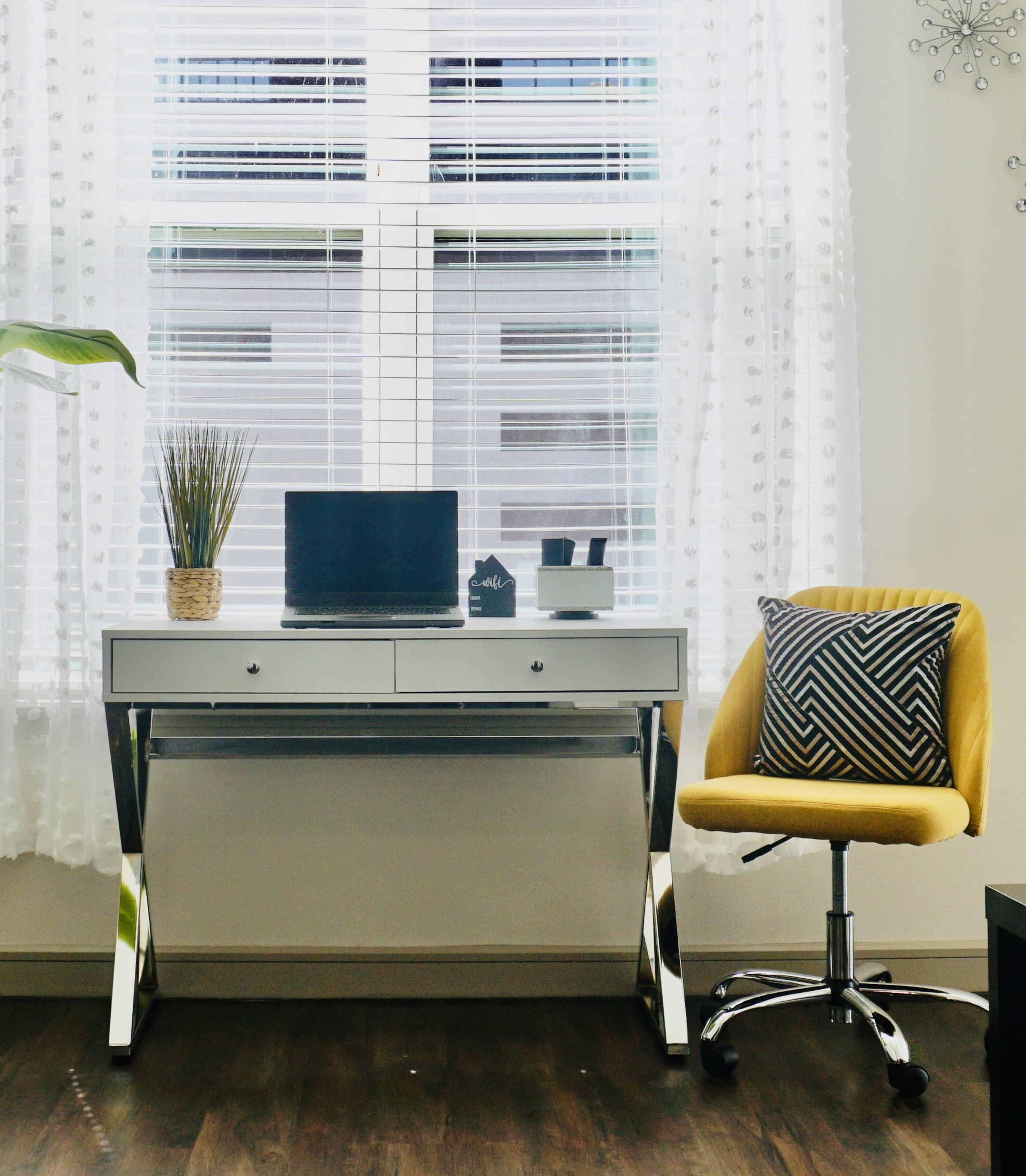 A modern office space features a sleek desk with a laptop, a potted plant, and a yellow swivel chair beside a window with sheer curtains.