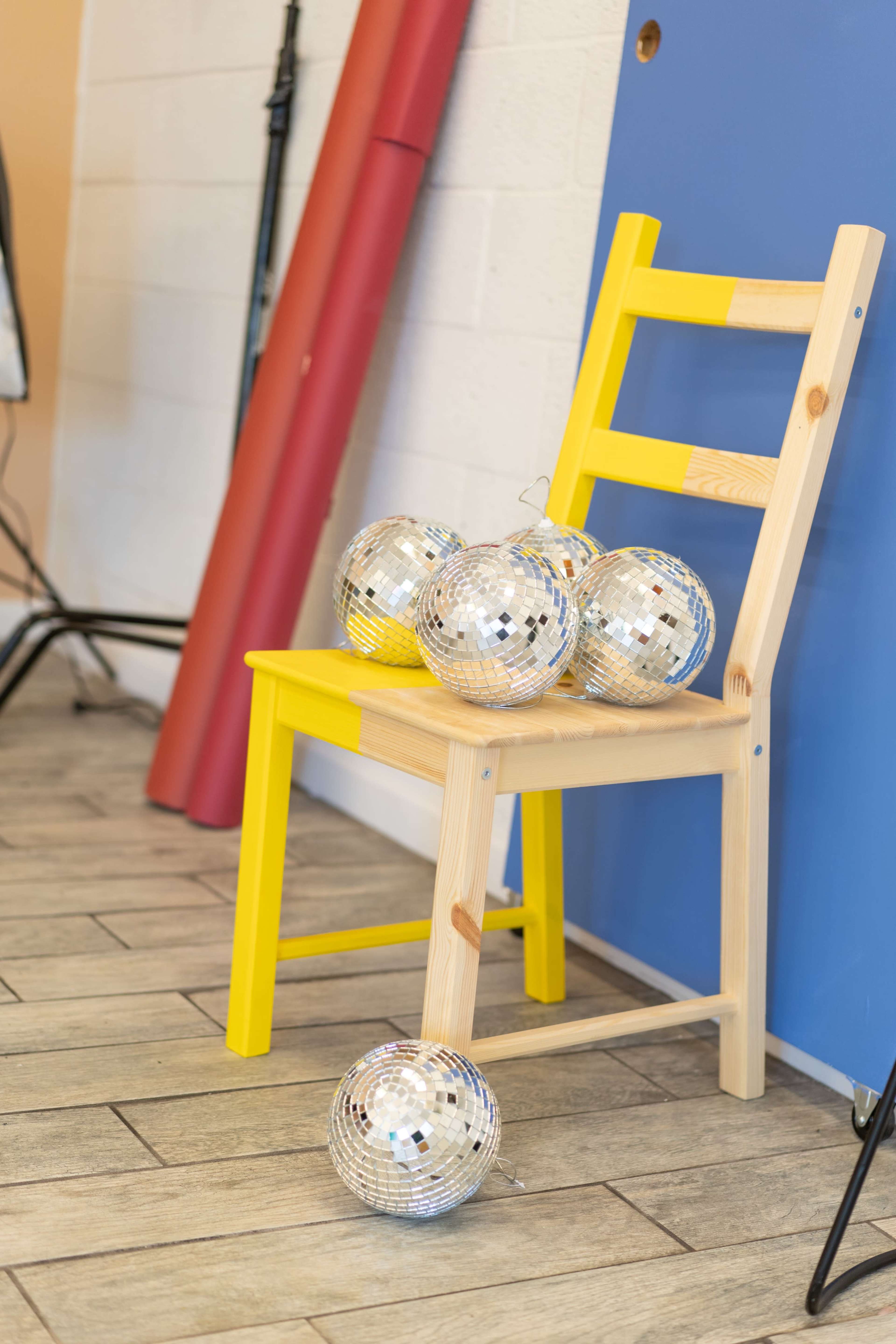 A yellow wooden chair holds several disco balls on a tiled floor near colored backdrops.