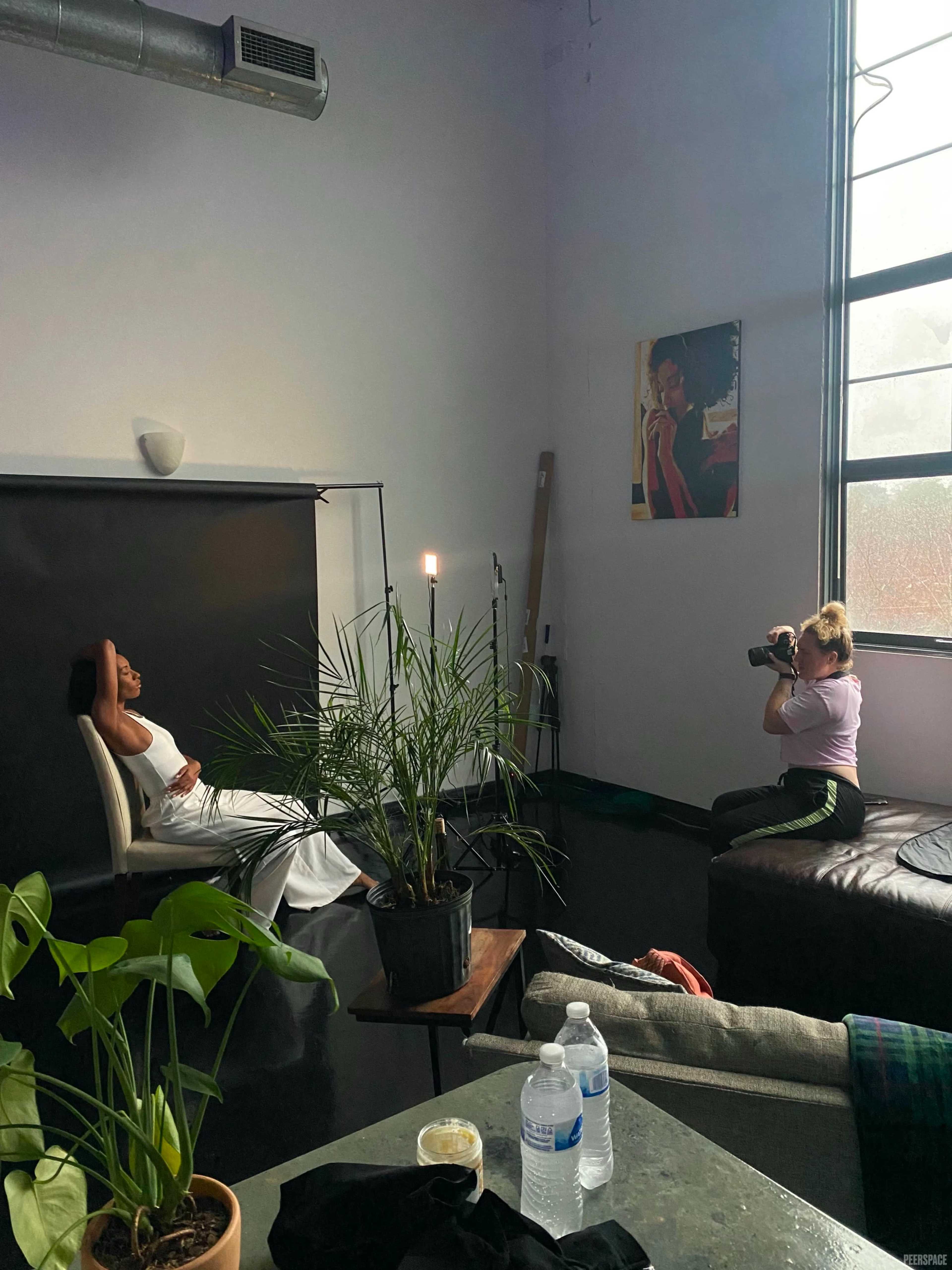 A woman in a white dress poses for a photographer in a studio with large windows and plants.