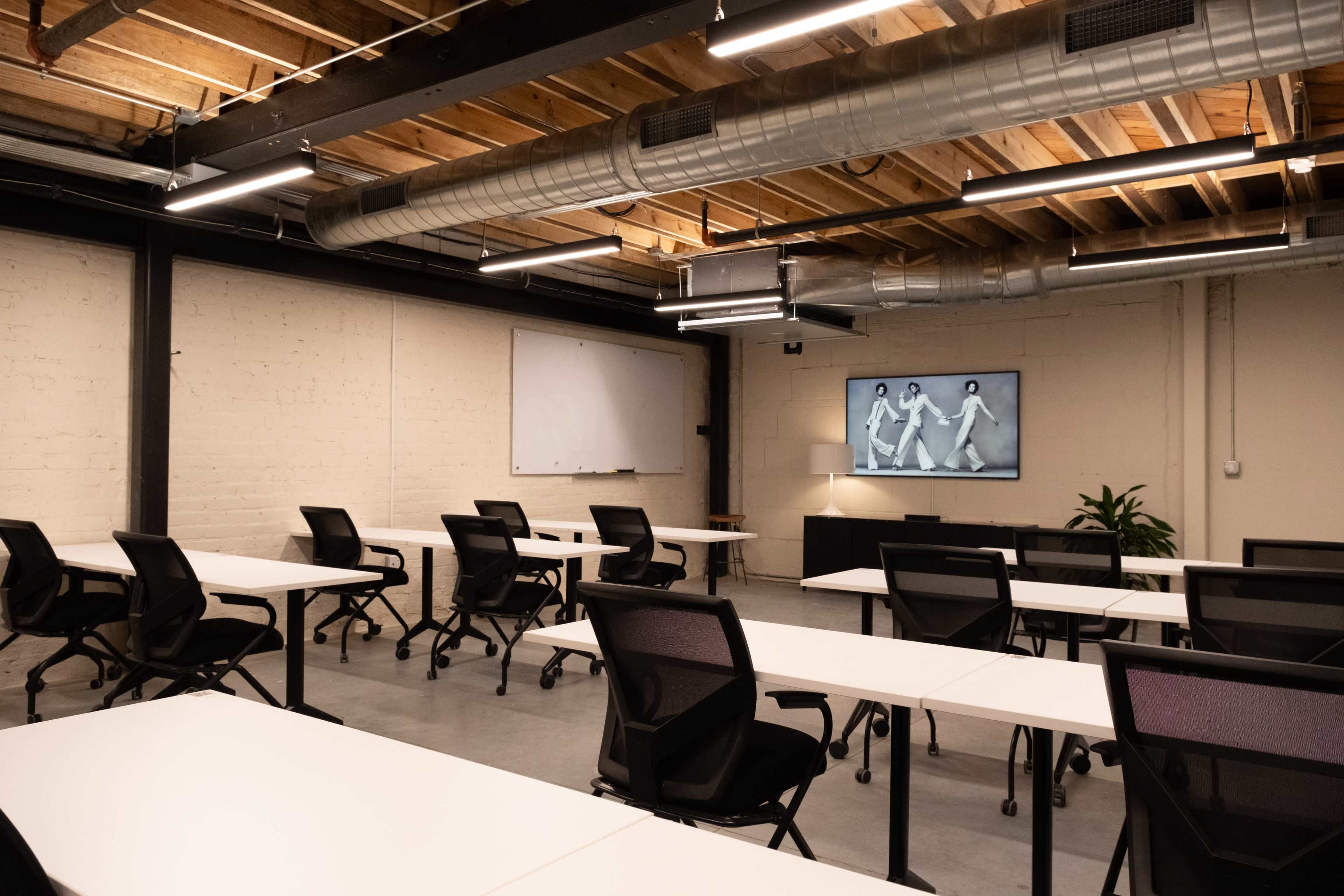 The image shows an empty training room with multiple rows of white tables and black chairs, featuring a wall-mounted screen displaying a monochrome image.