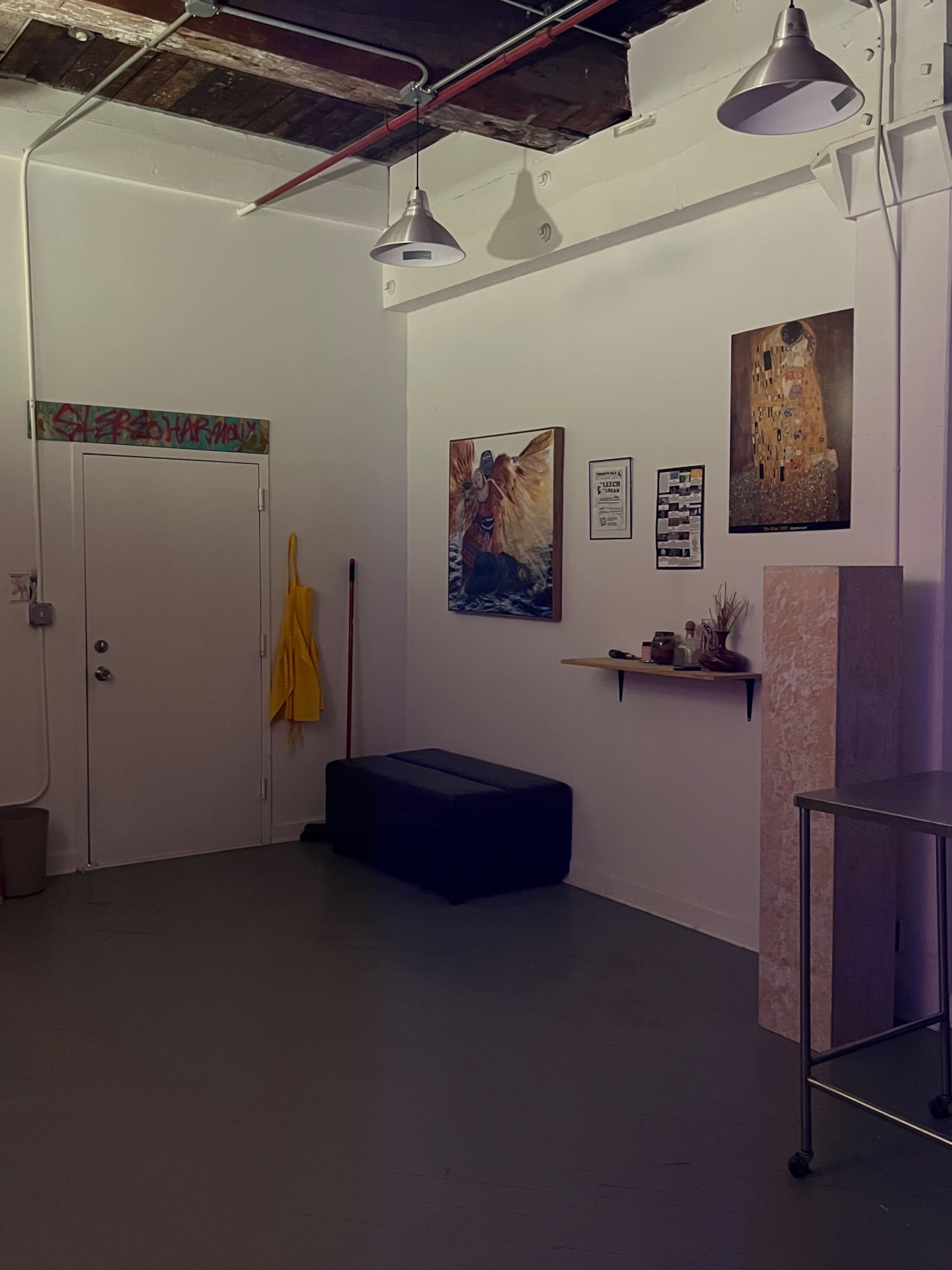 The image shows a sparsely decorated room with a dark floor, a yellow coat hanging on the wall, and various artworks and shelves arranged around the space.