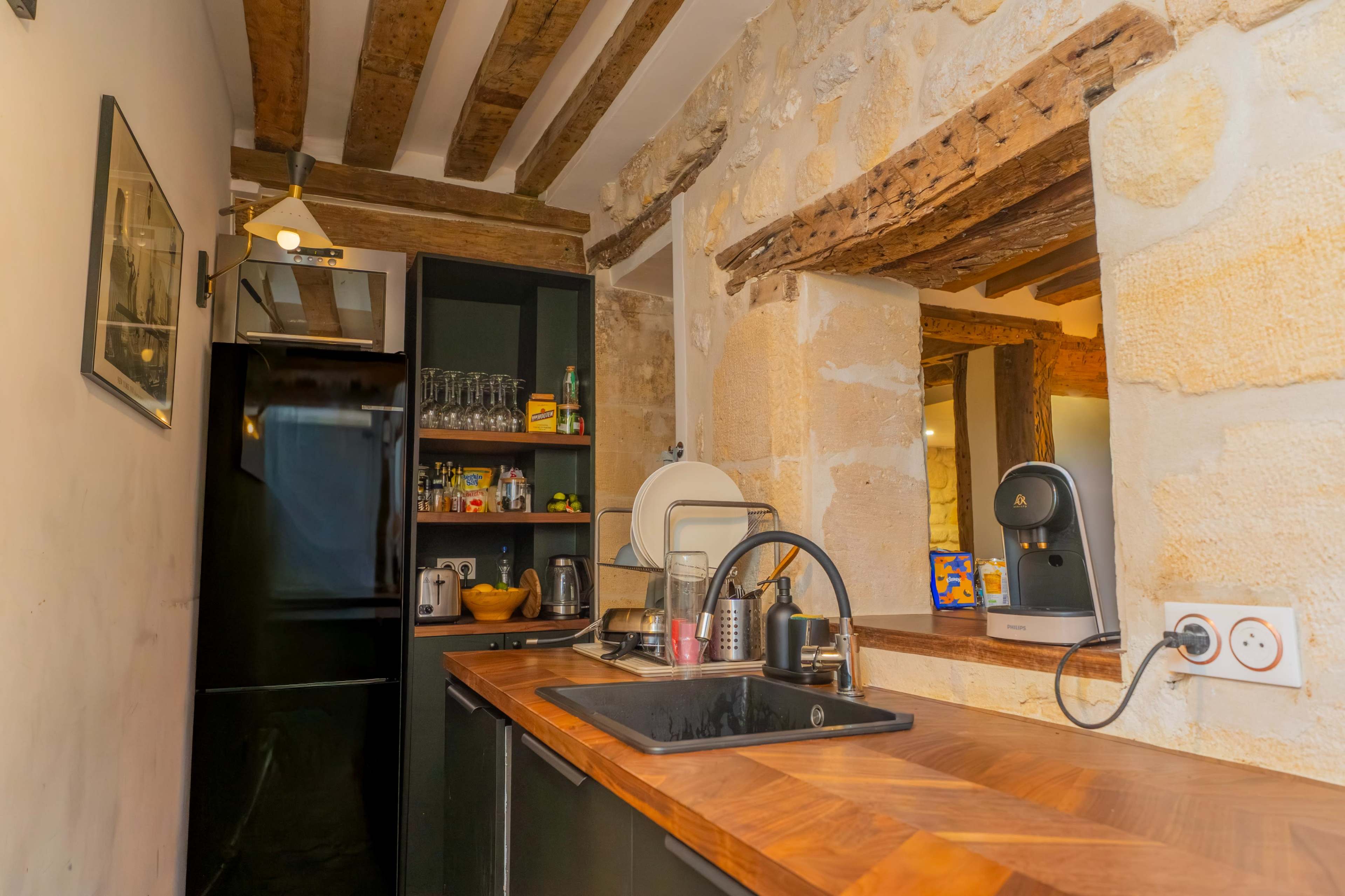 The image shows a small kitchen featuring dark cabinetry, wooden countertops, and exposed stone walls, along with a coffee machine and various kitchen appliances.