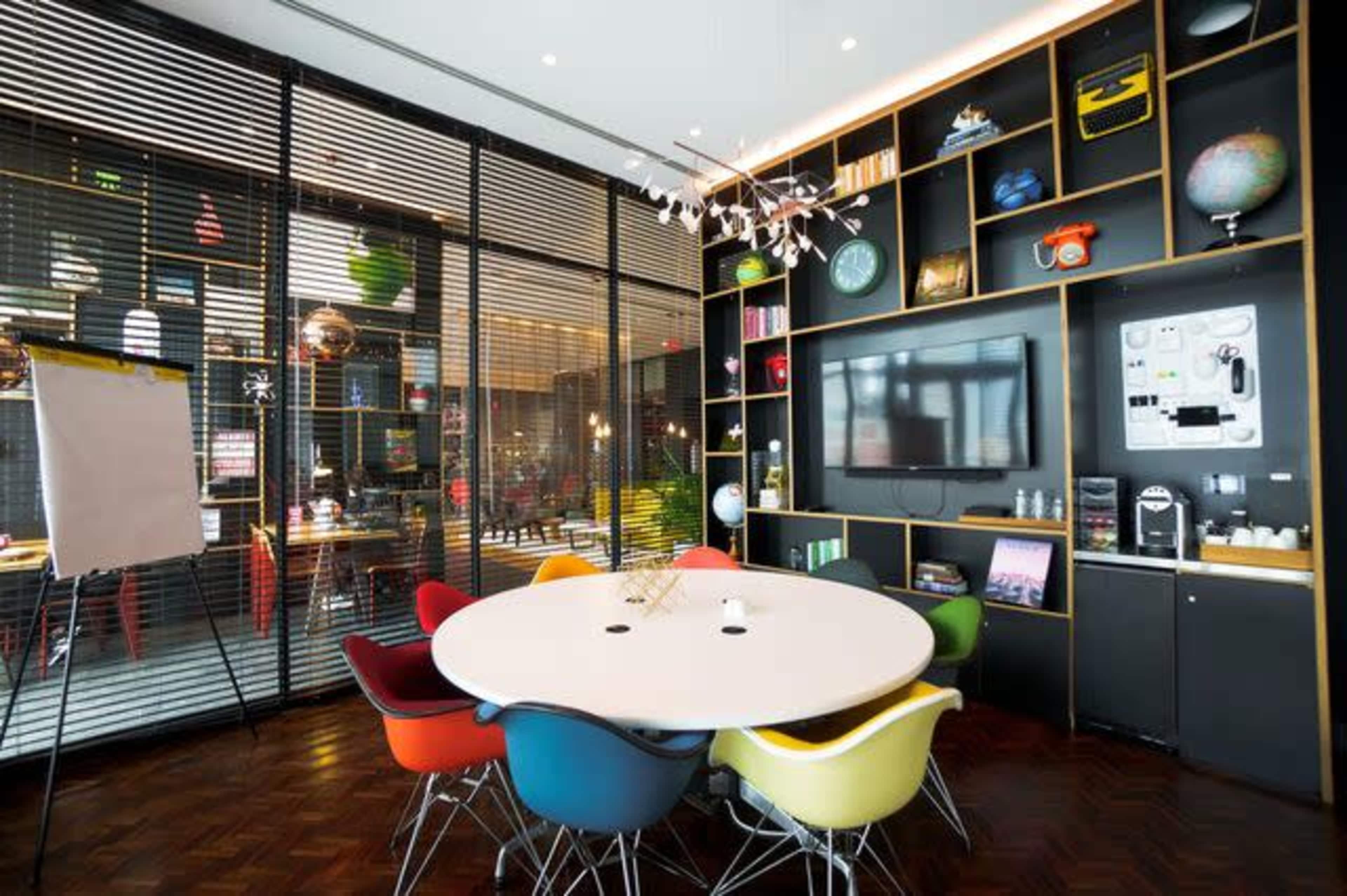 A modern conference room features a round table surrounded by colorful chairs, with a wall-mounted television and shelves displaying decorative items.