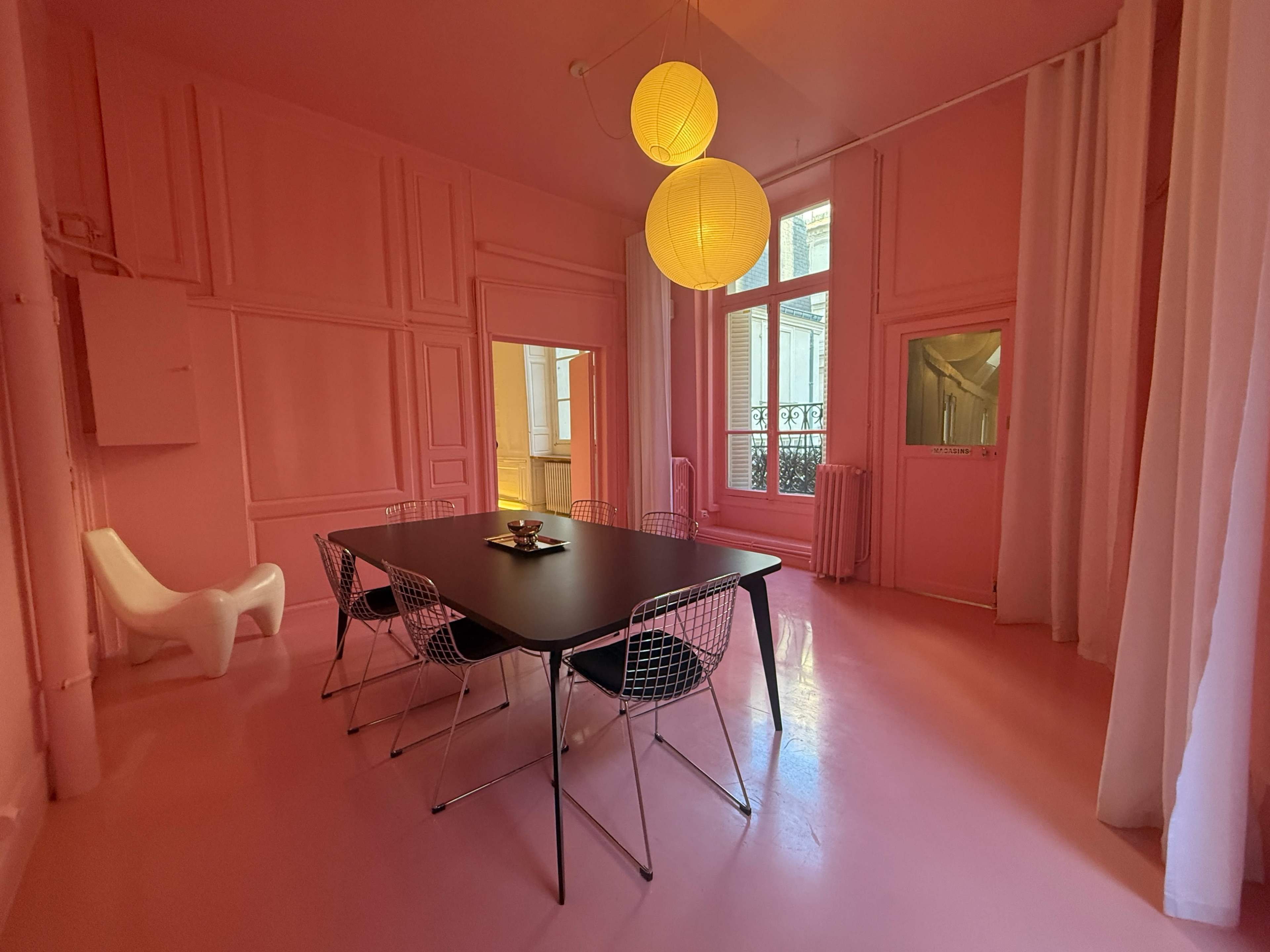 The image shows a pink room with a black dining table surrounded by six chairs, illuminated by two large pendant lamps.