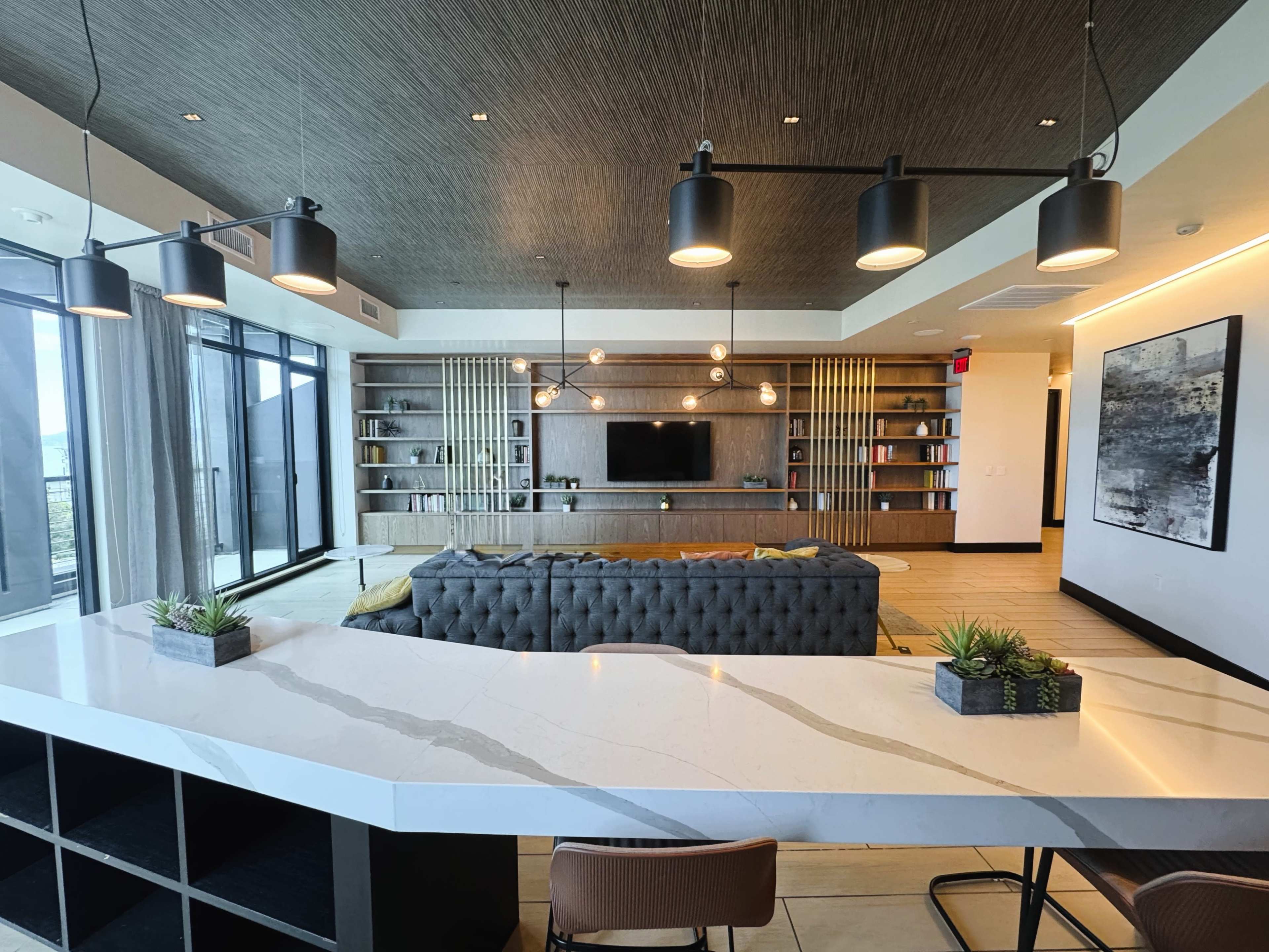 The image shows a modern lounge area featuring a gray tufted sofa, a wall-mounted television, bookshelves, and large windows providing natural light, with a kitchen countertop in the foreground.