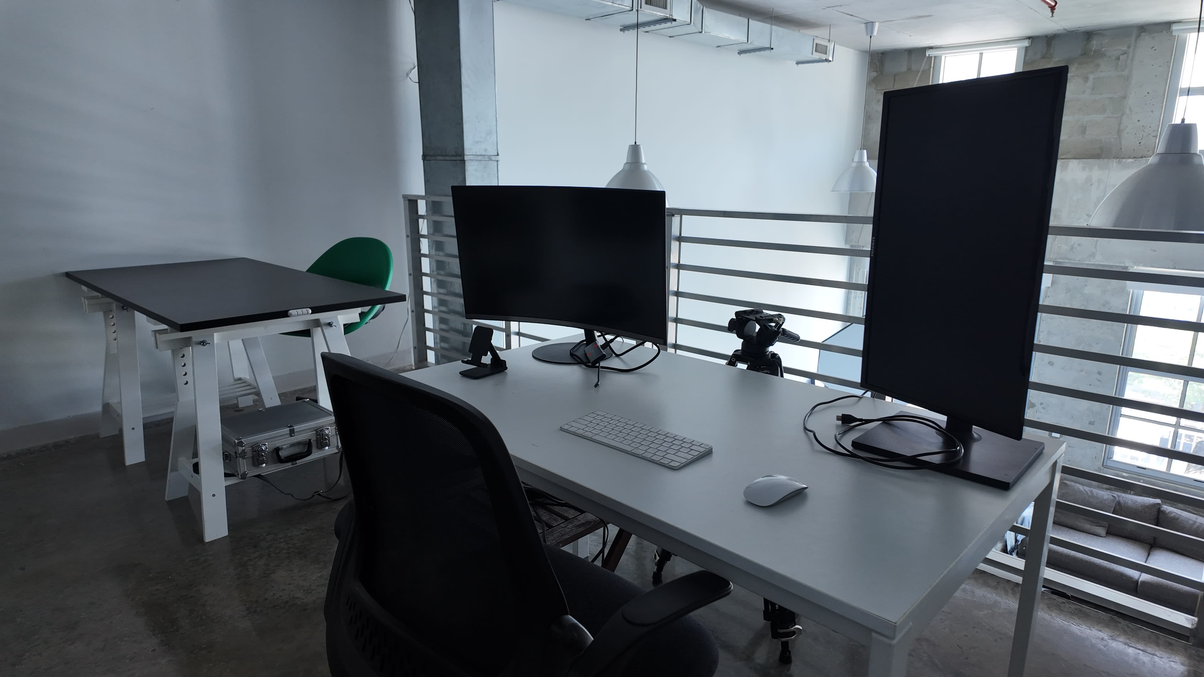 The image shows a modern workspace featuring a desk with two monitors, a keyboard, a mouse, a camera, and a chair, all set in a bright area with large windows.
