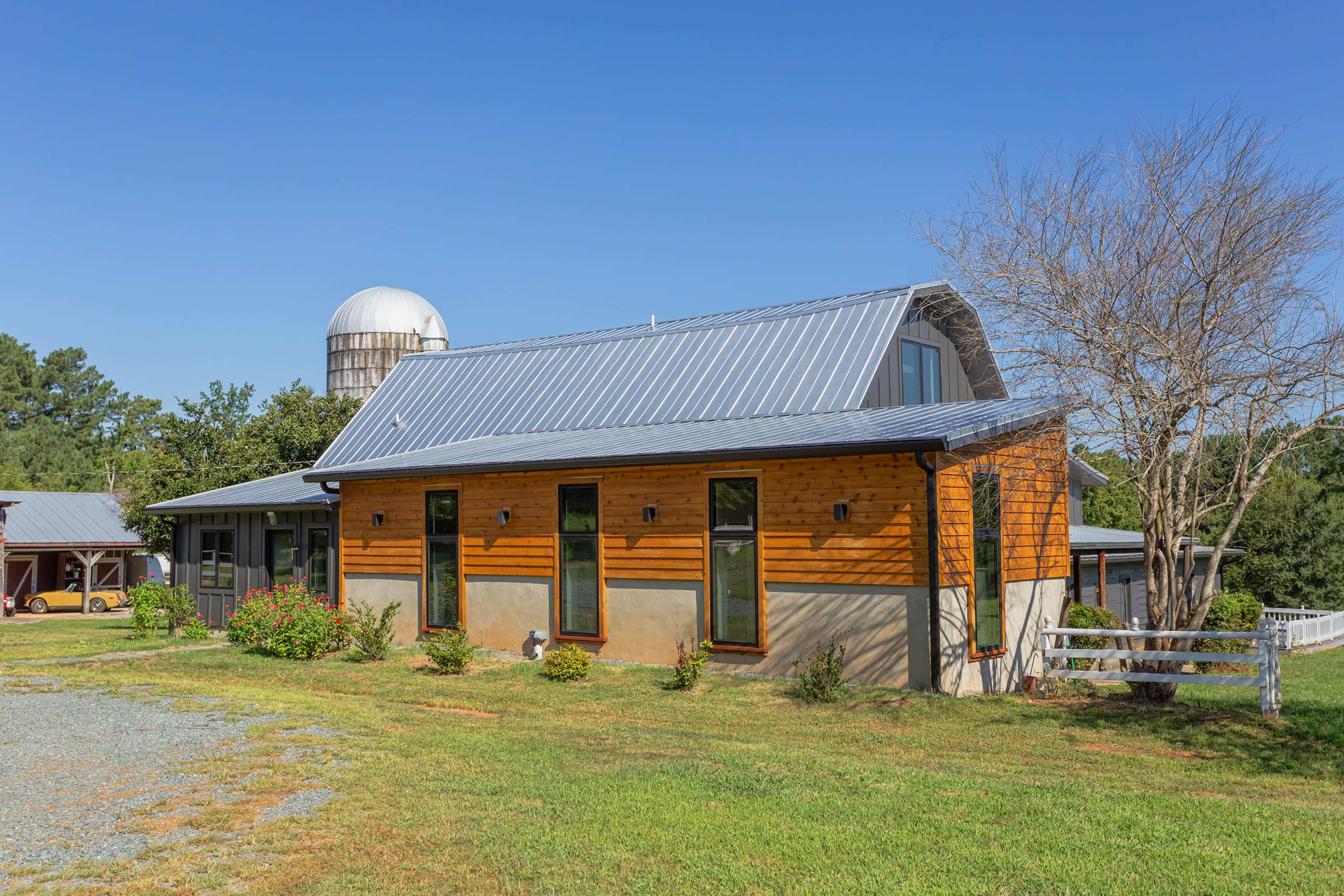 A modern farmhouse with a metal roof and wooden accents is set against a clear blue sky, surrounded by green grass and a silos in the background.