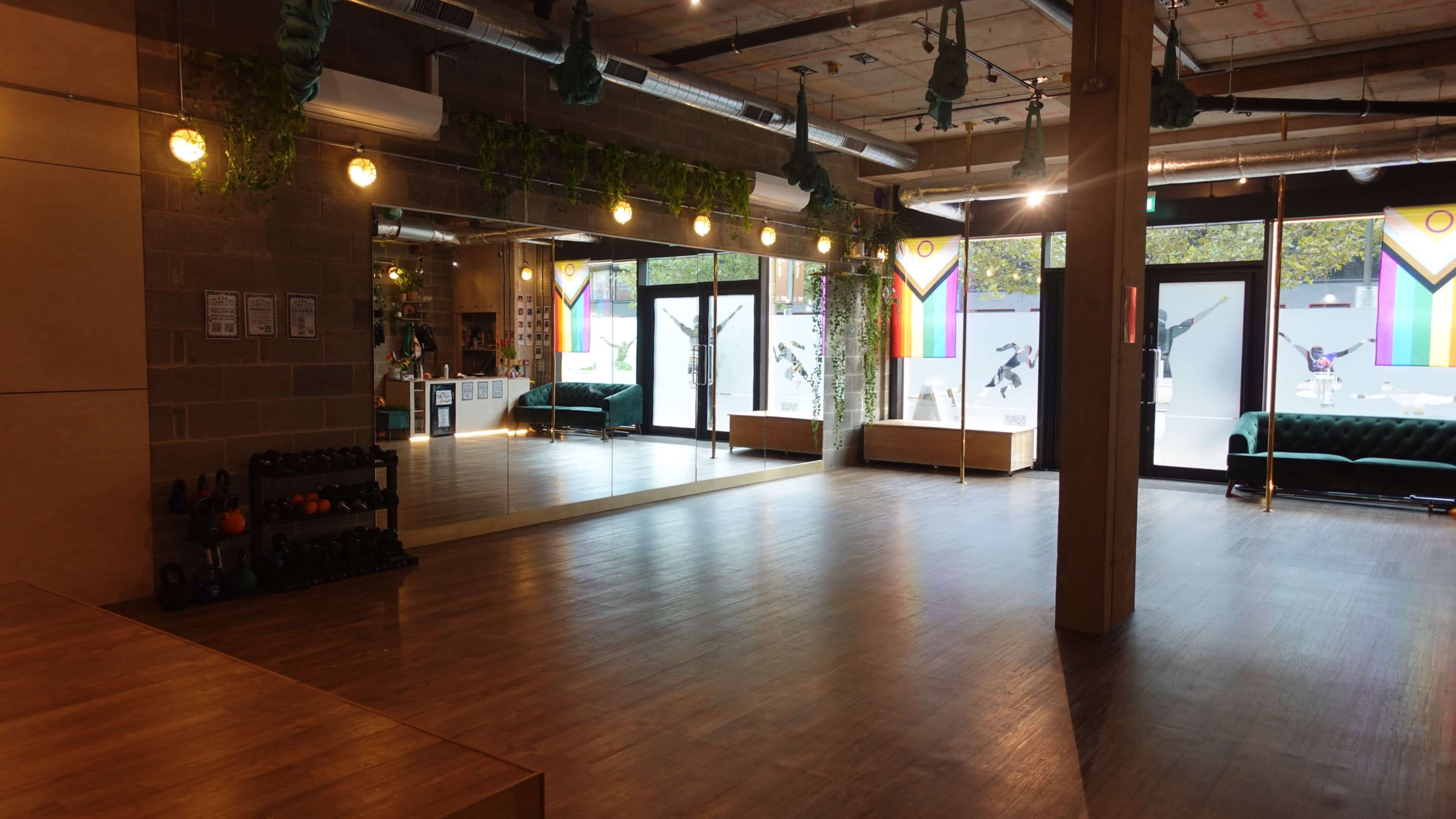 The image shows a spacious interior of a fitness studio featuring wooden flooring, large mirrors, and bright natural light coming through the front windows.