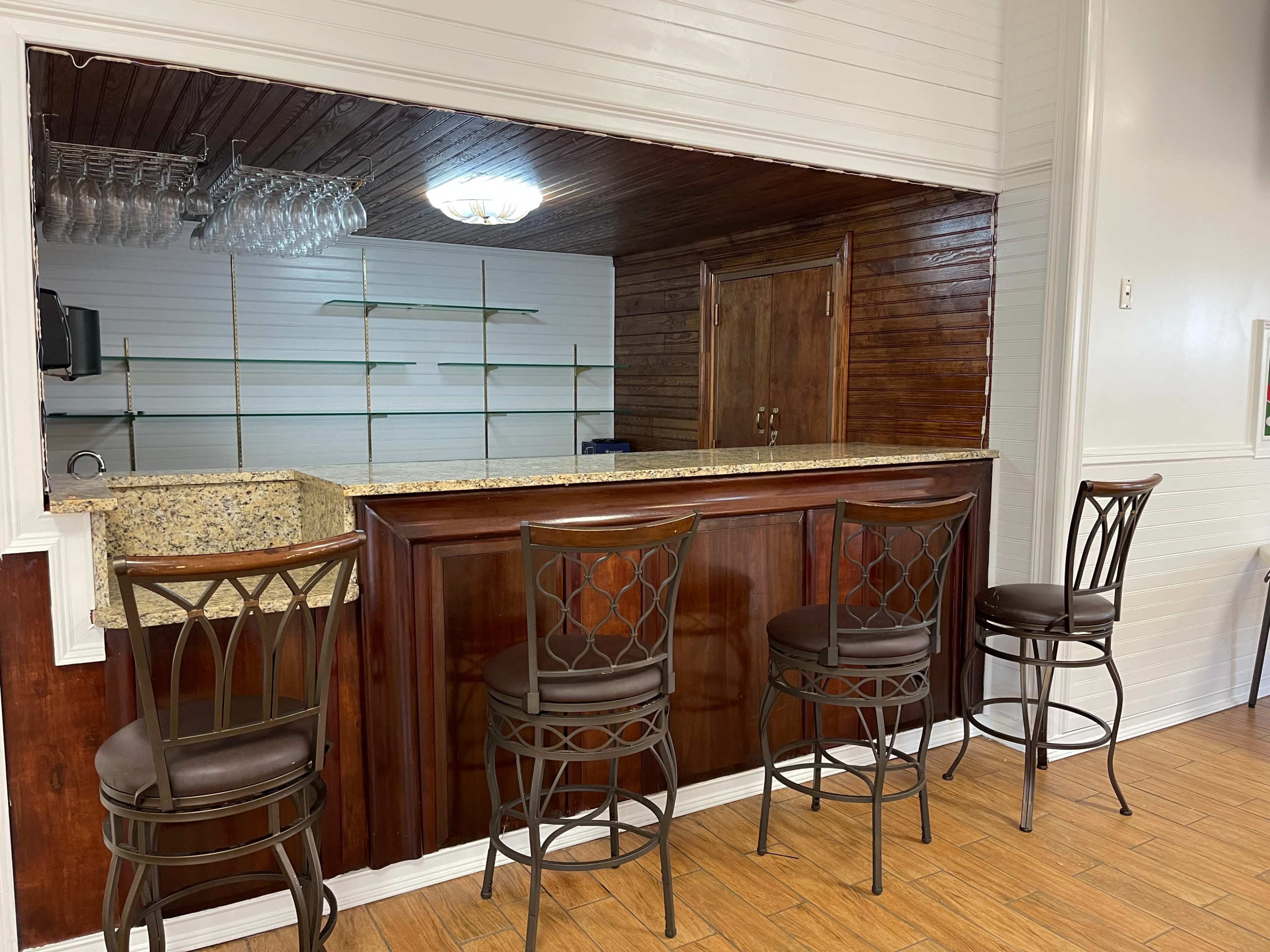 The image shows a wooden bar with four metal stools in front of a mirrored backsplash and a light fixture above.