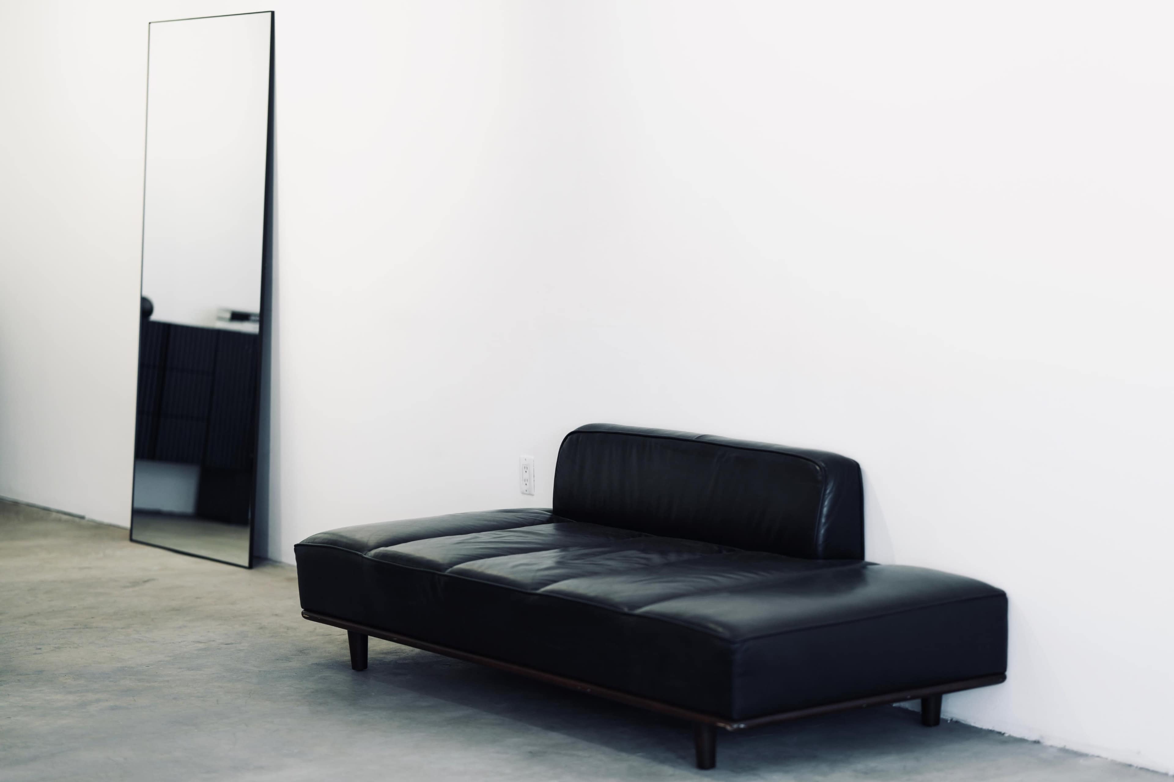 The image shows a modern black sofa placed next to a tall, rectangular mirror against a plain white wall in a minimalist room.