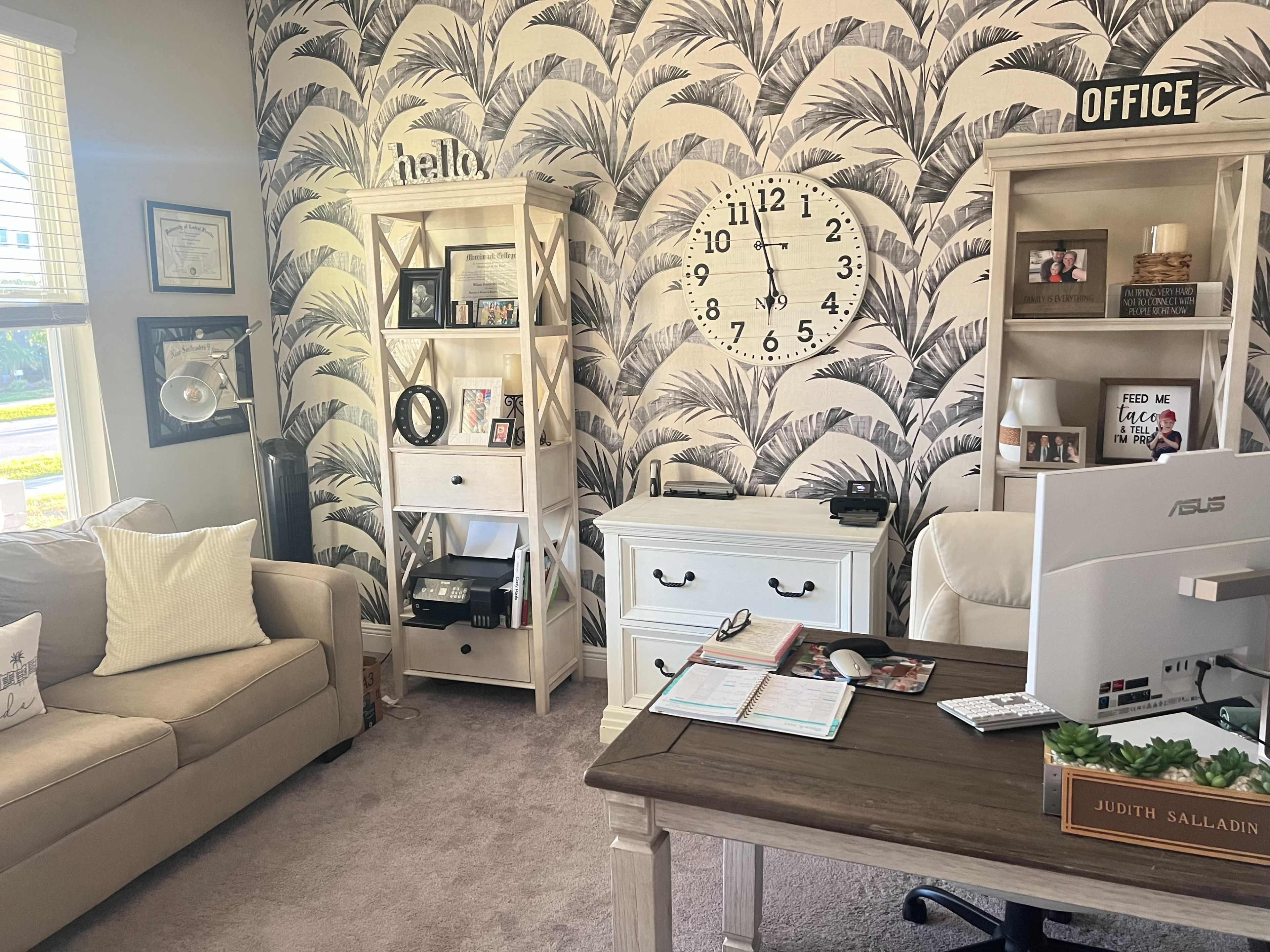 A home office featuring a desk with a computer, bookshelves, and a decorative wall with a tropical leaf pattern.