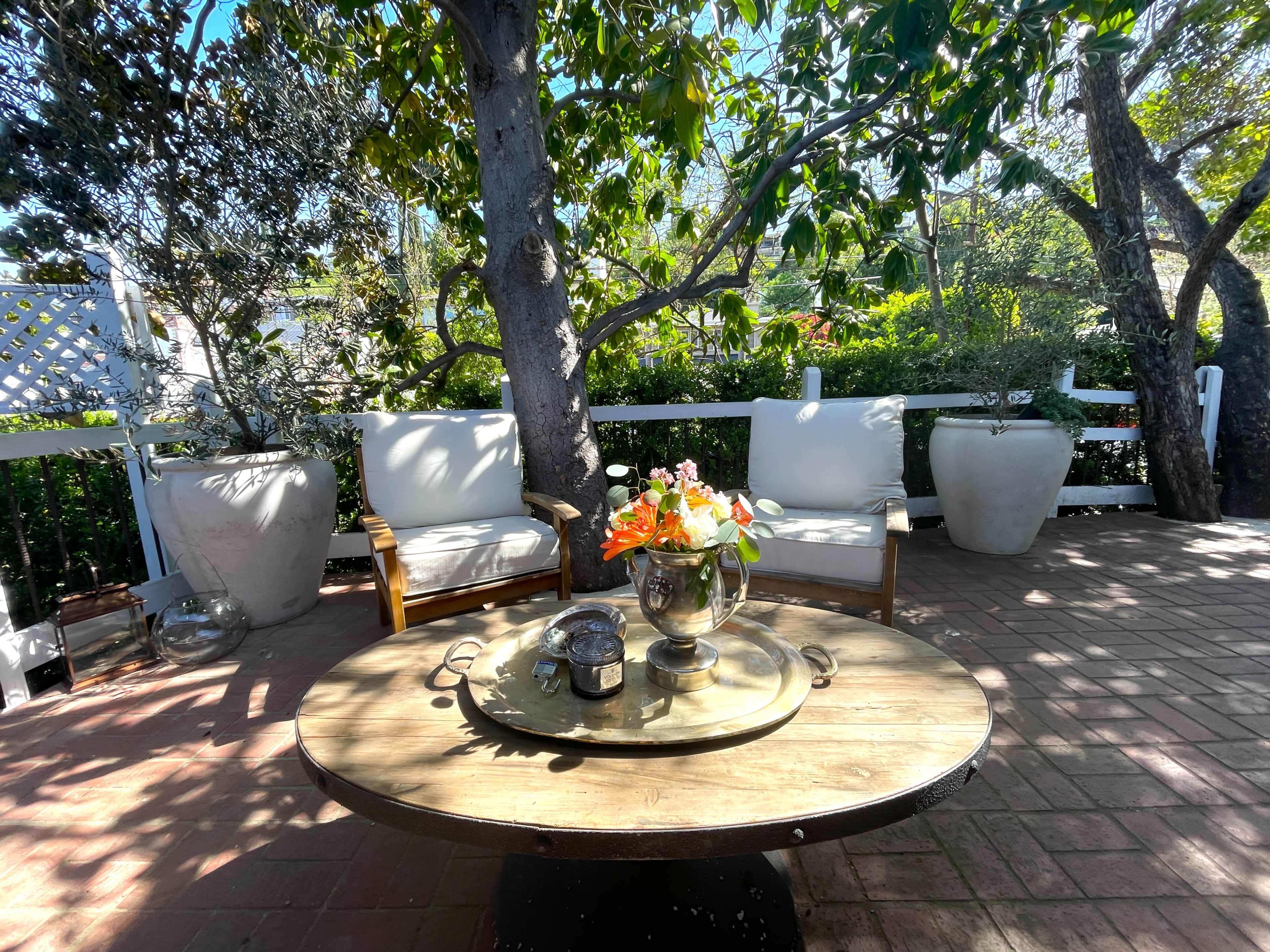 A wooden table with a floral arrangement sits in an outdoor seating area surrounded by greenery and shaded by trees.