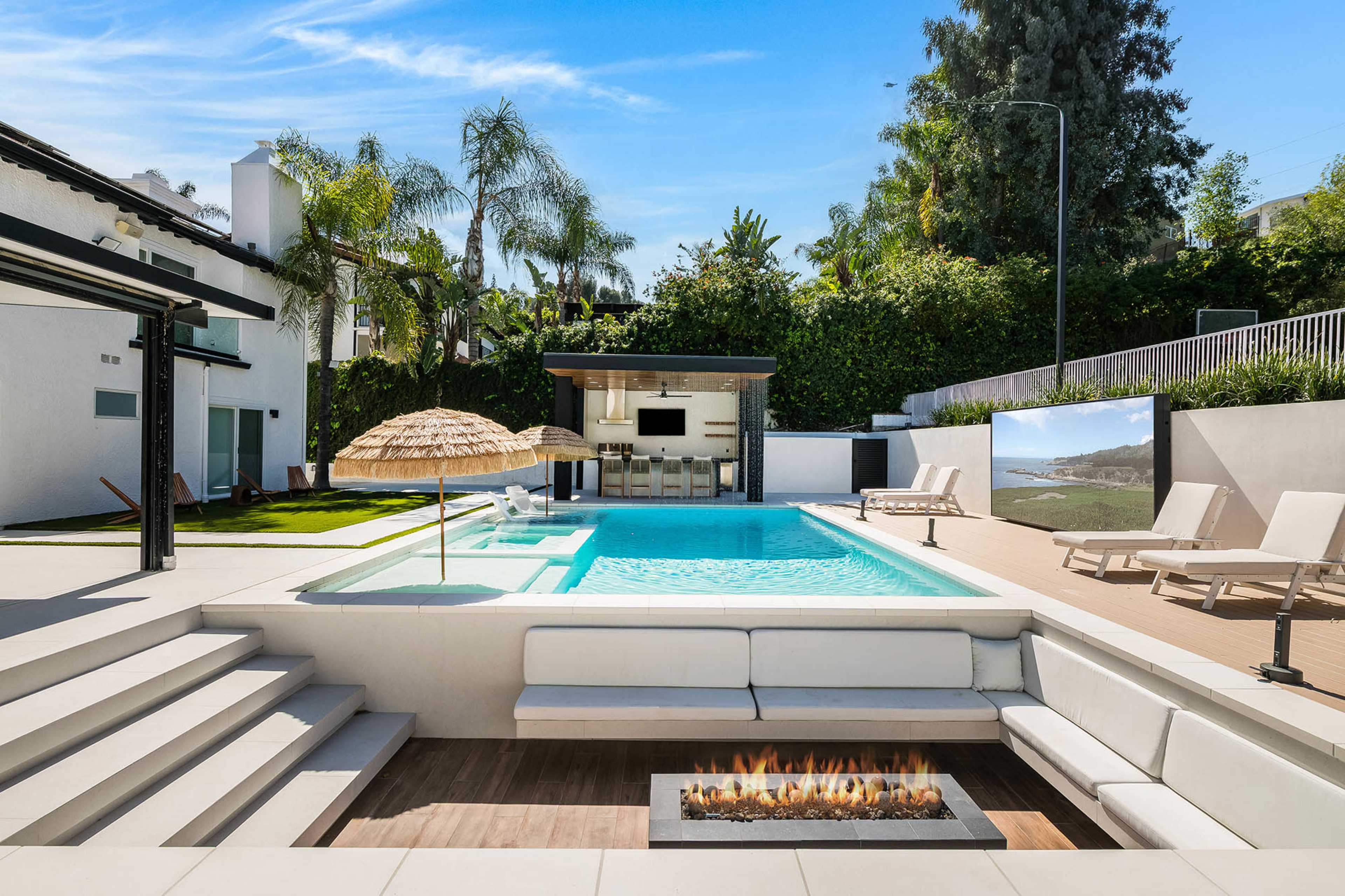 A modern outdoor space features a swimming pool, lounge chairs, a fire pit, and a shaded area with an umbrella.