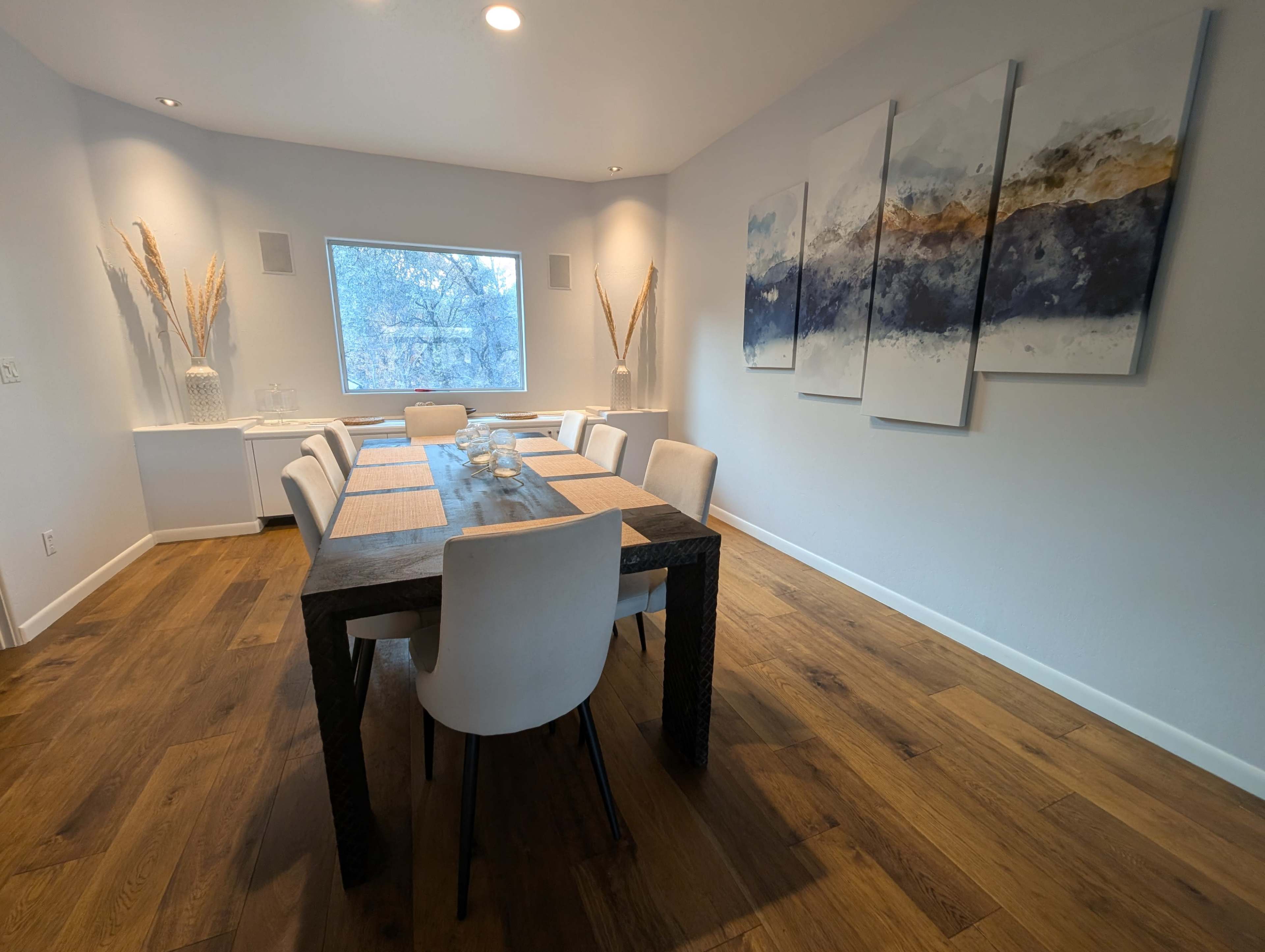 A modern dining room features a long table set with placemats, surrounded by white chairs, under a window with a view of trees.