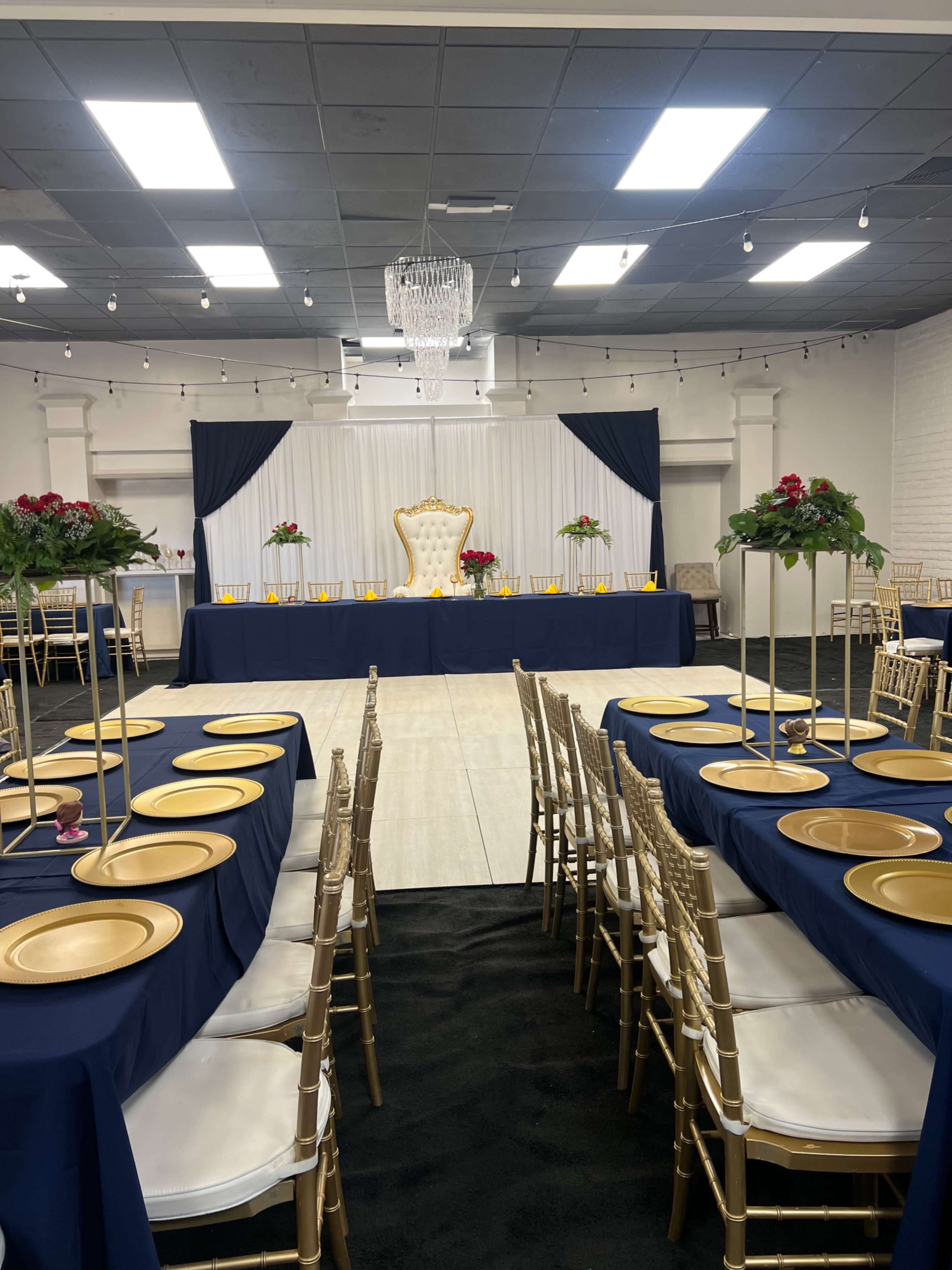 A banquet hall set up with gold and navy table settings, featuring a raised backdrop with a throne and floral arrangements at the front.