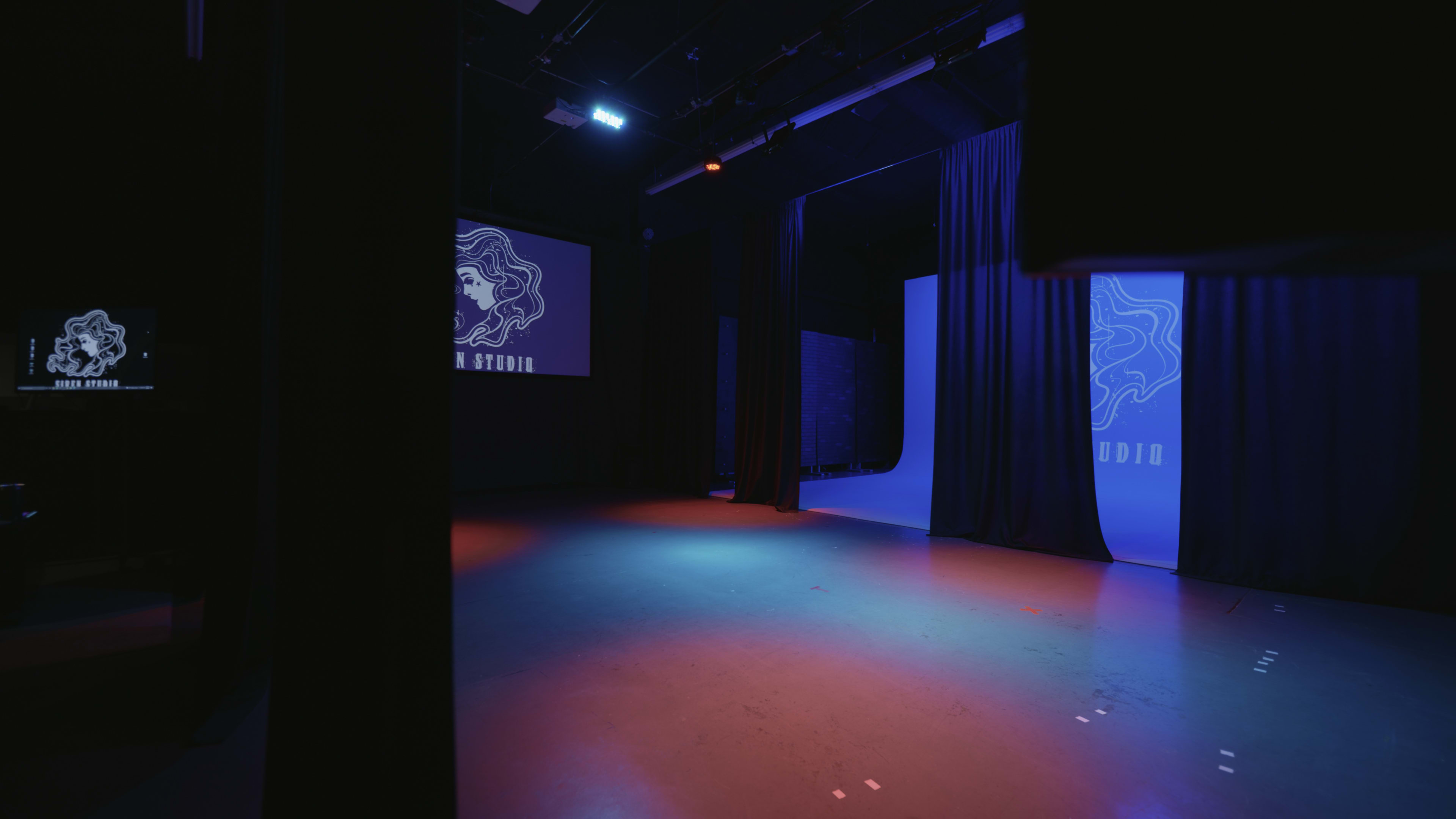 A dimly lit performance studio with blue and purple lighting, featuring a stage area partially obscured by black curtains.
