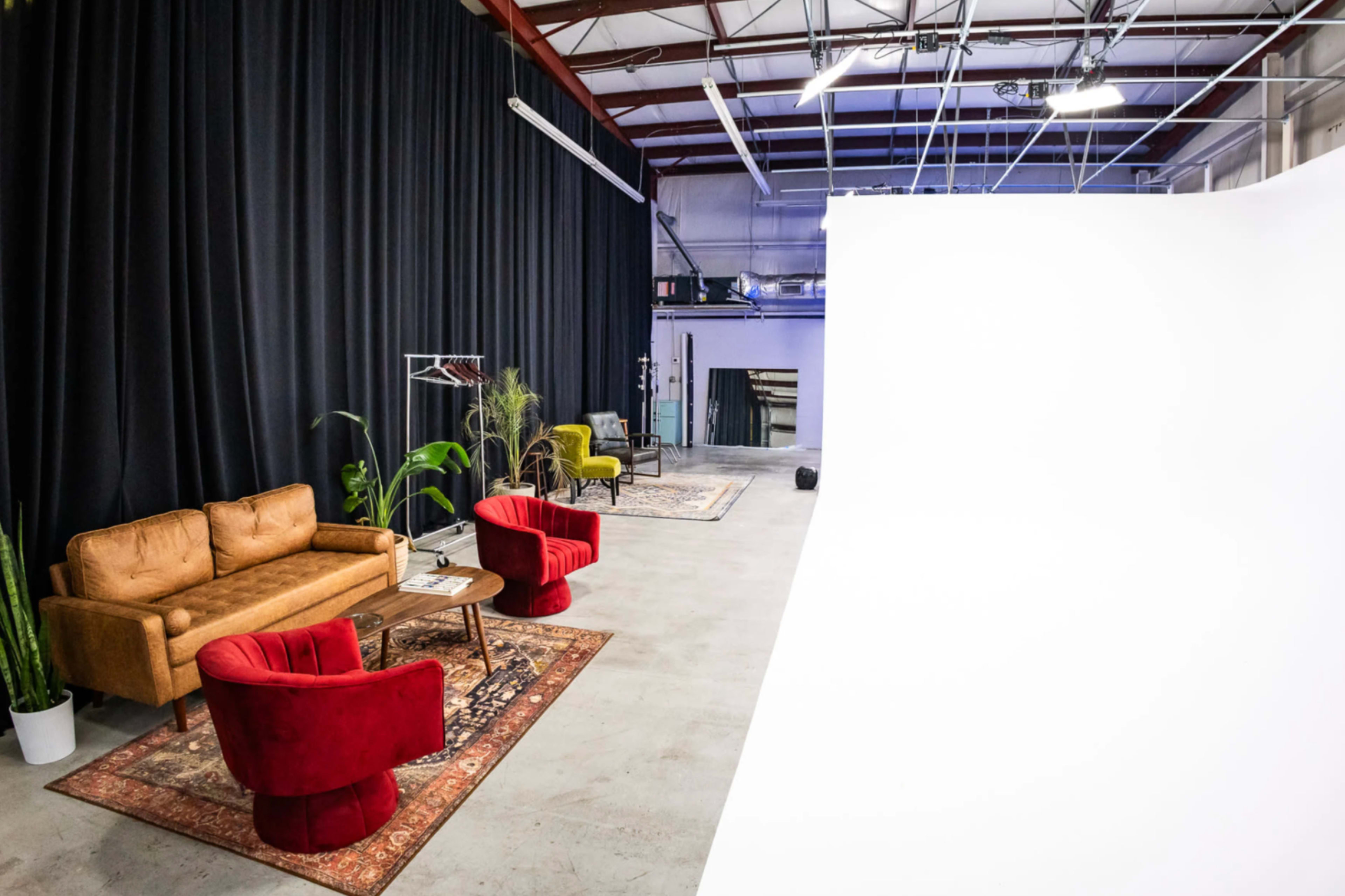 The image shows a spacious studio with a seating area featuring a brown sofa and red armchairs, accompanied by a small table and backdrop.