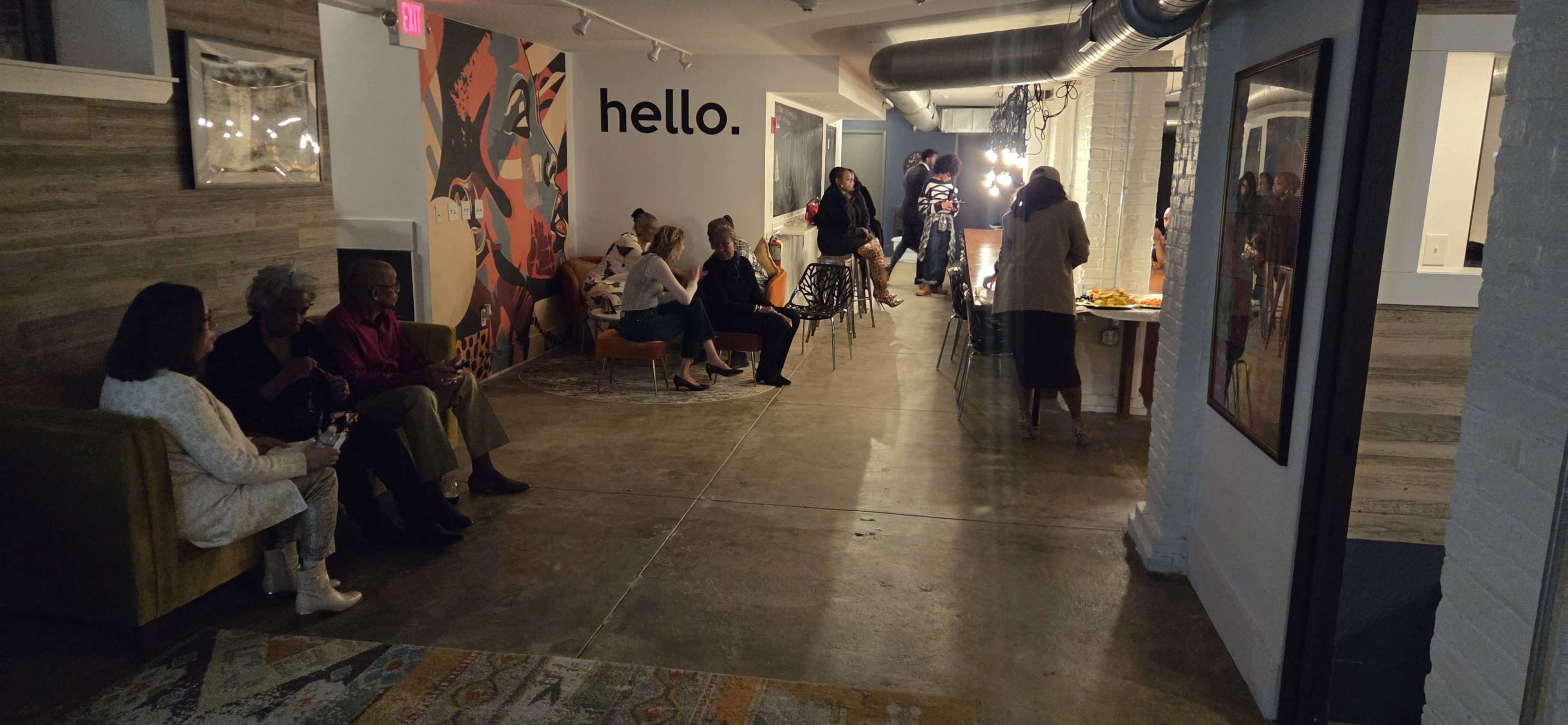 The image shows a modern indoor space with a mix of people socializing, seated on couches, and engaging in conversation near a wall that features a mural with the word "hello."