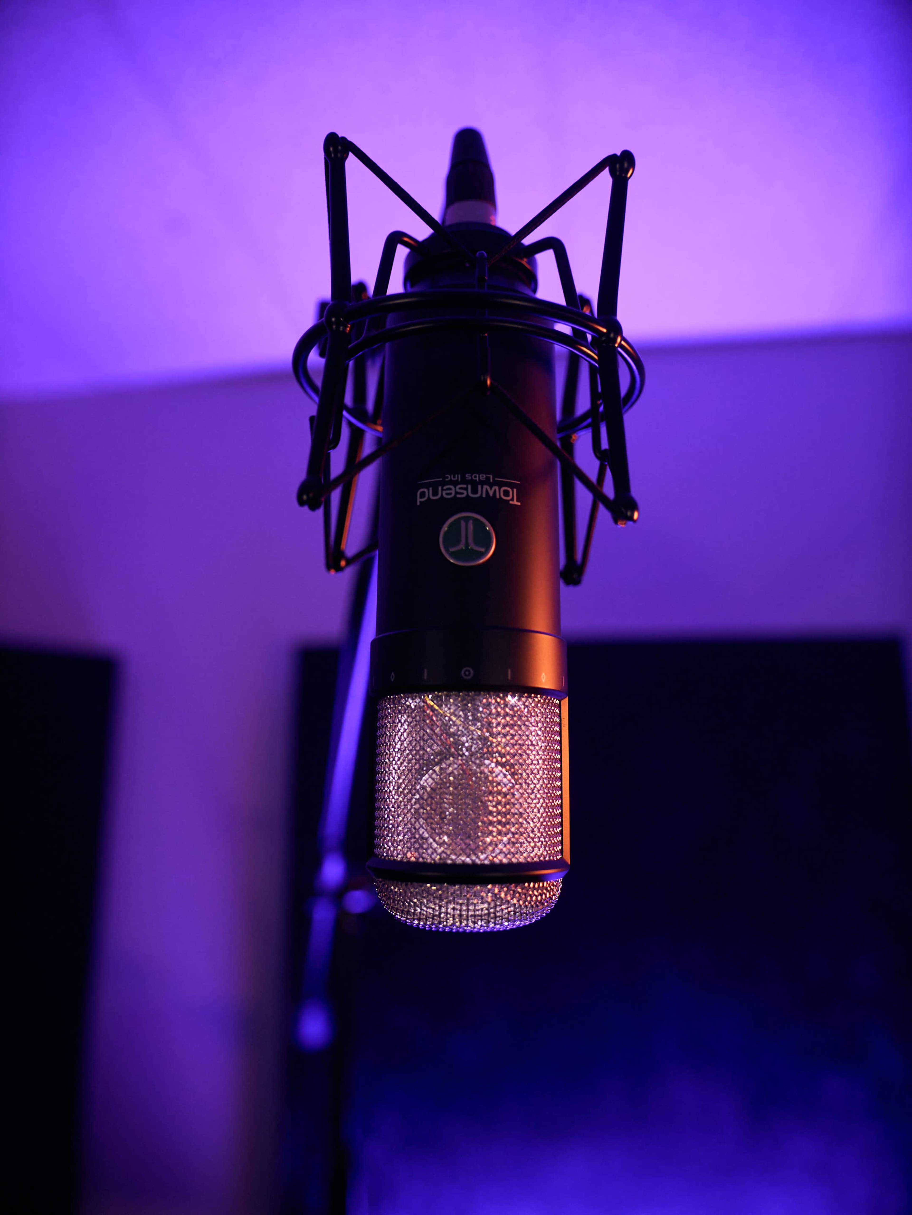 A black studio microphone hangs from a ceiling mount in a dimly lit space with purple lighting.