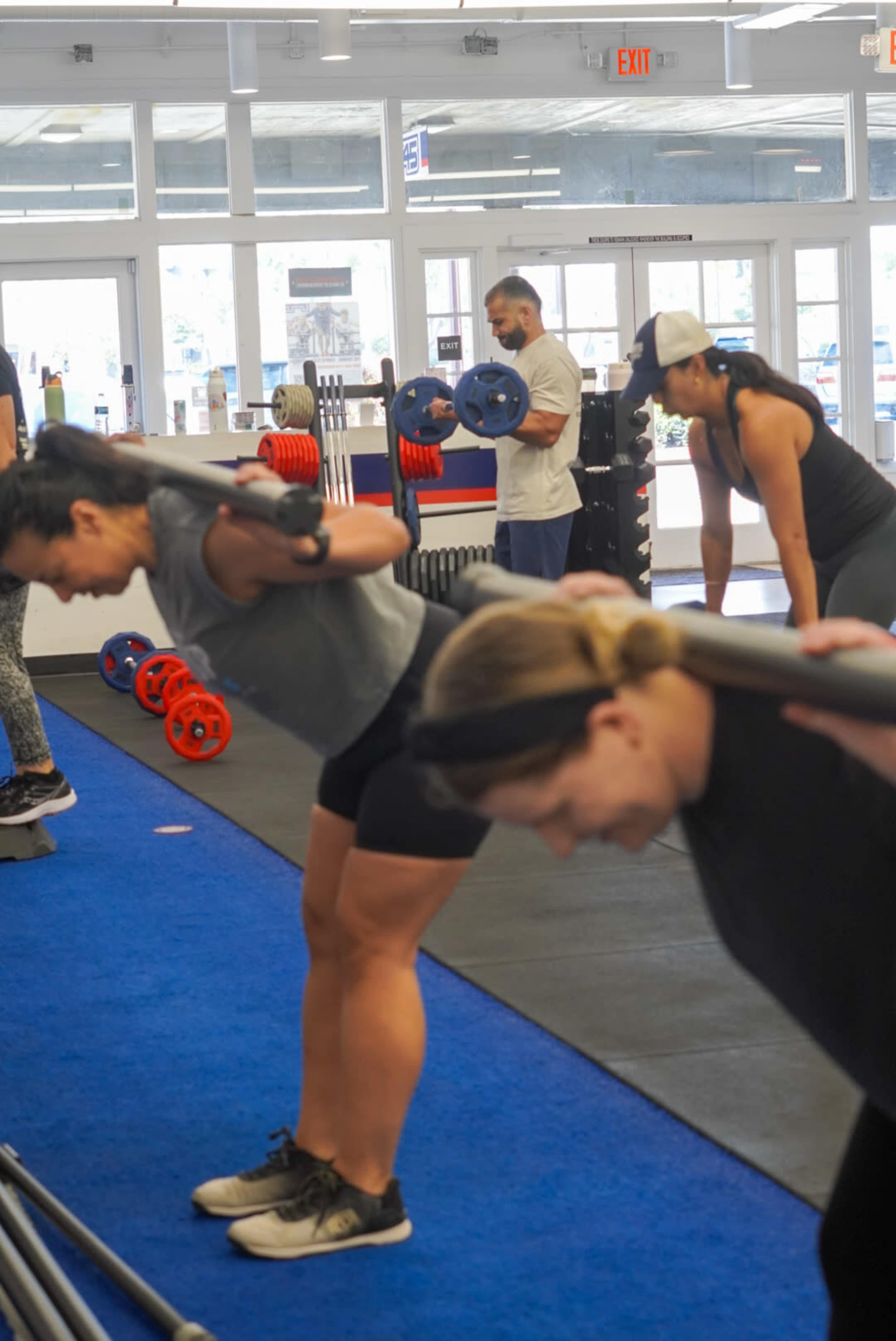 Several individuals are performing bent-over rows with weights in a gym setting, with exercise equipment and weights visible in the background.