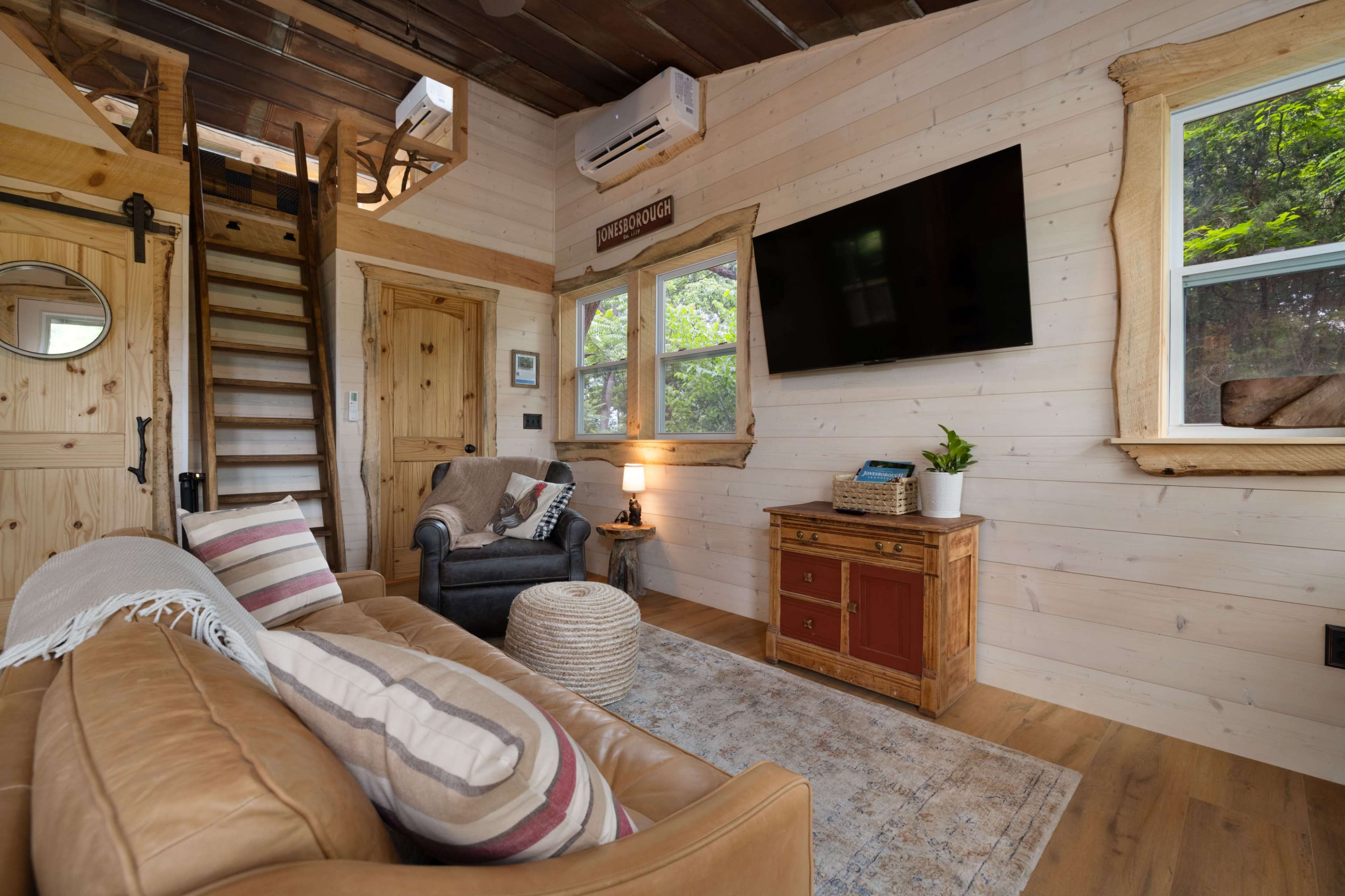 The image shows a cozy interior of a wooden cabin with a living area featuring a leather sofa, a chair, a television mounted on the wall, and stairs leading to a loft.
