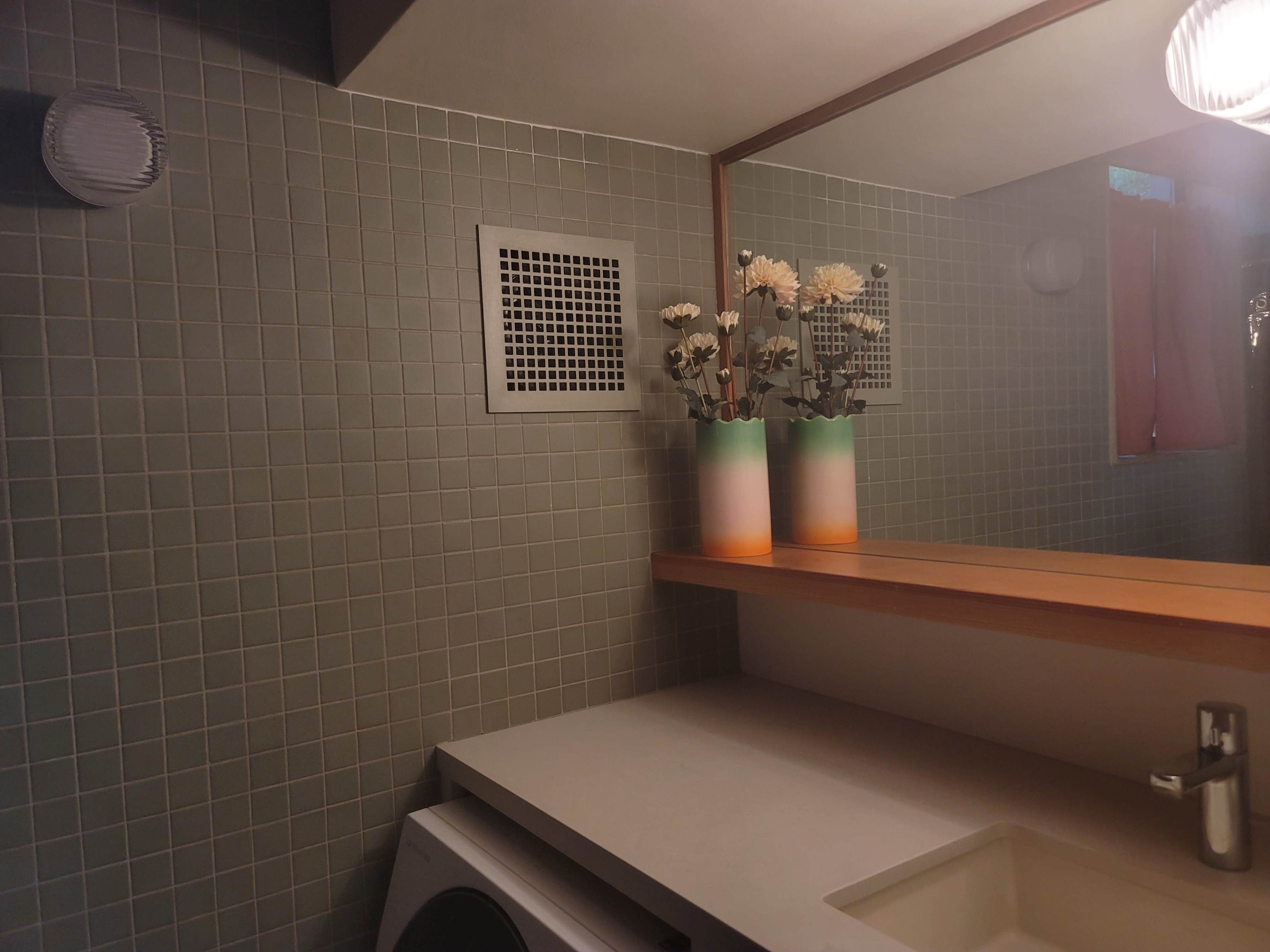 The image shows a modern bathroom with green tiled walls, a mirrored shelf above a countertop, and two vases with flowers on display.