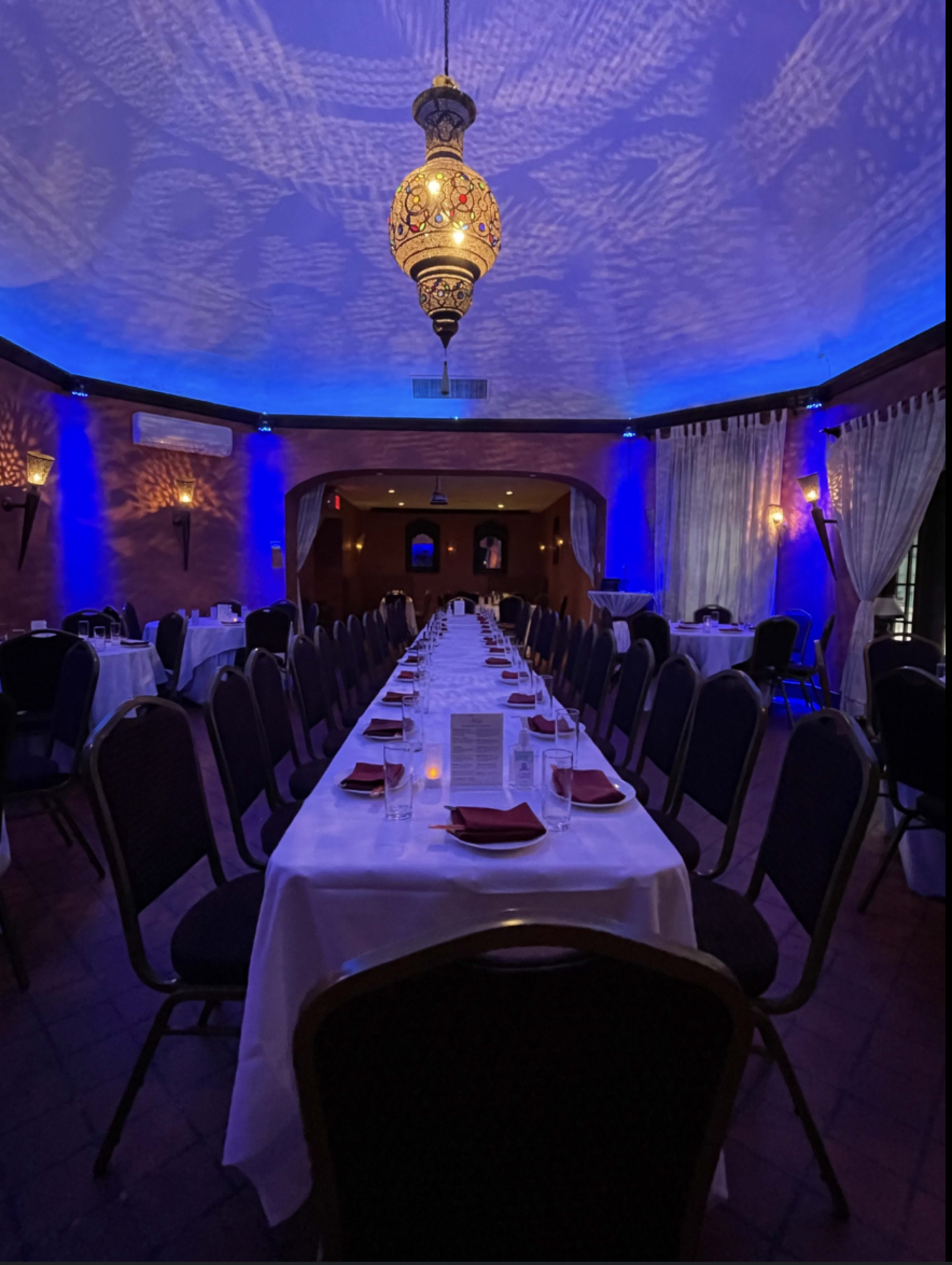 The image shows a long dining table set with white tablecloths and red napkins in a dimly lit restaurant, featuring blue-lit walls and a decorative ceiling fixture.