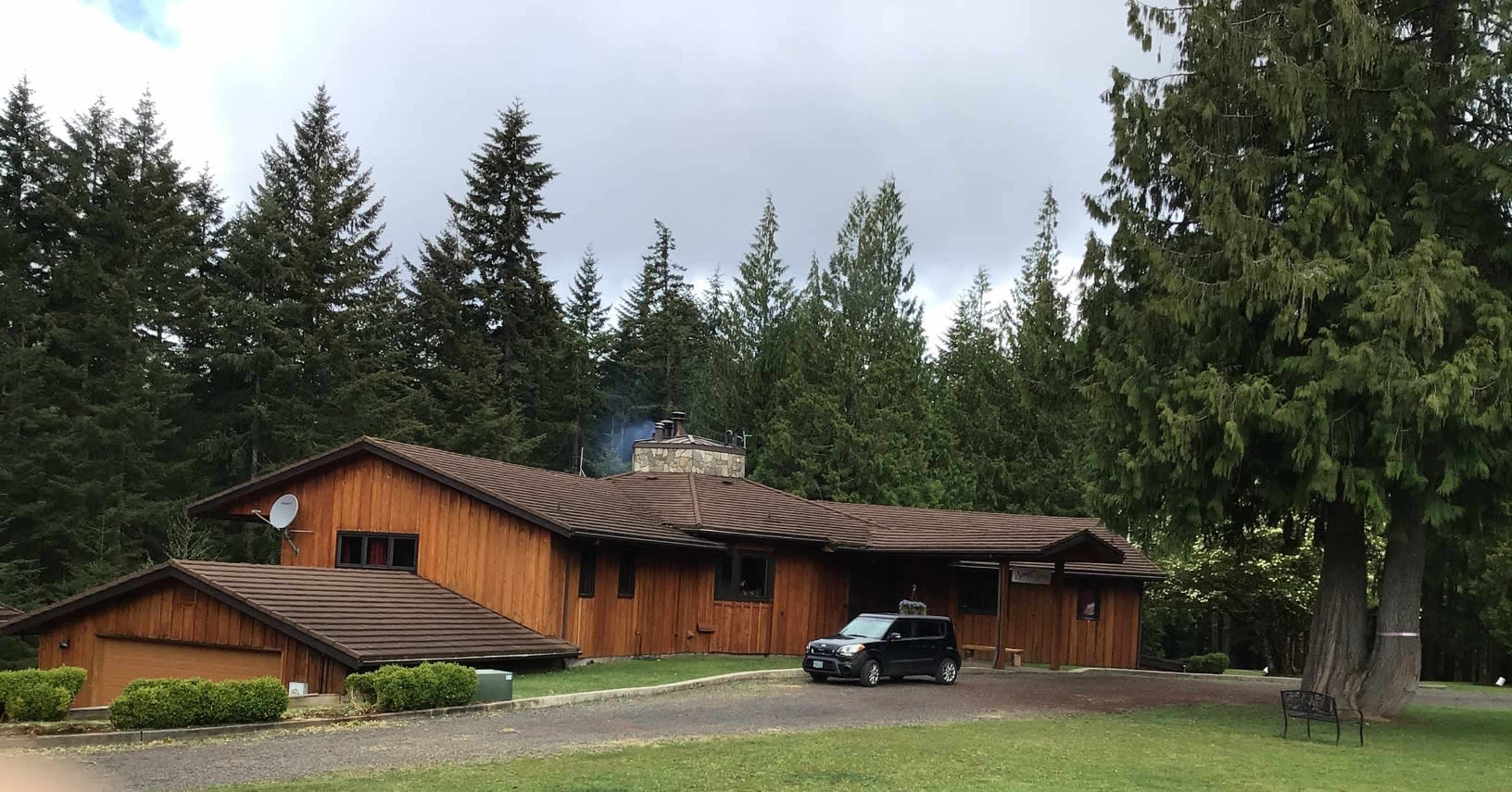 A wooden house with a stone chimney sits amidst tall trees, with a small car parked in front on a gravel drive.