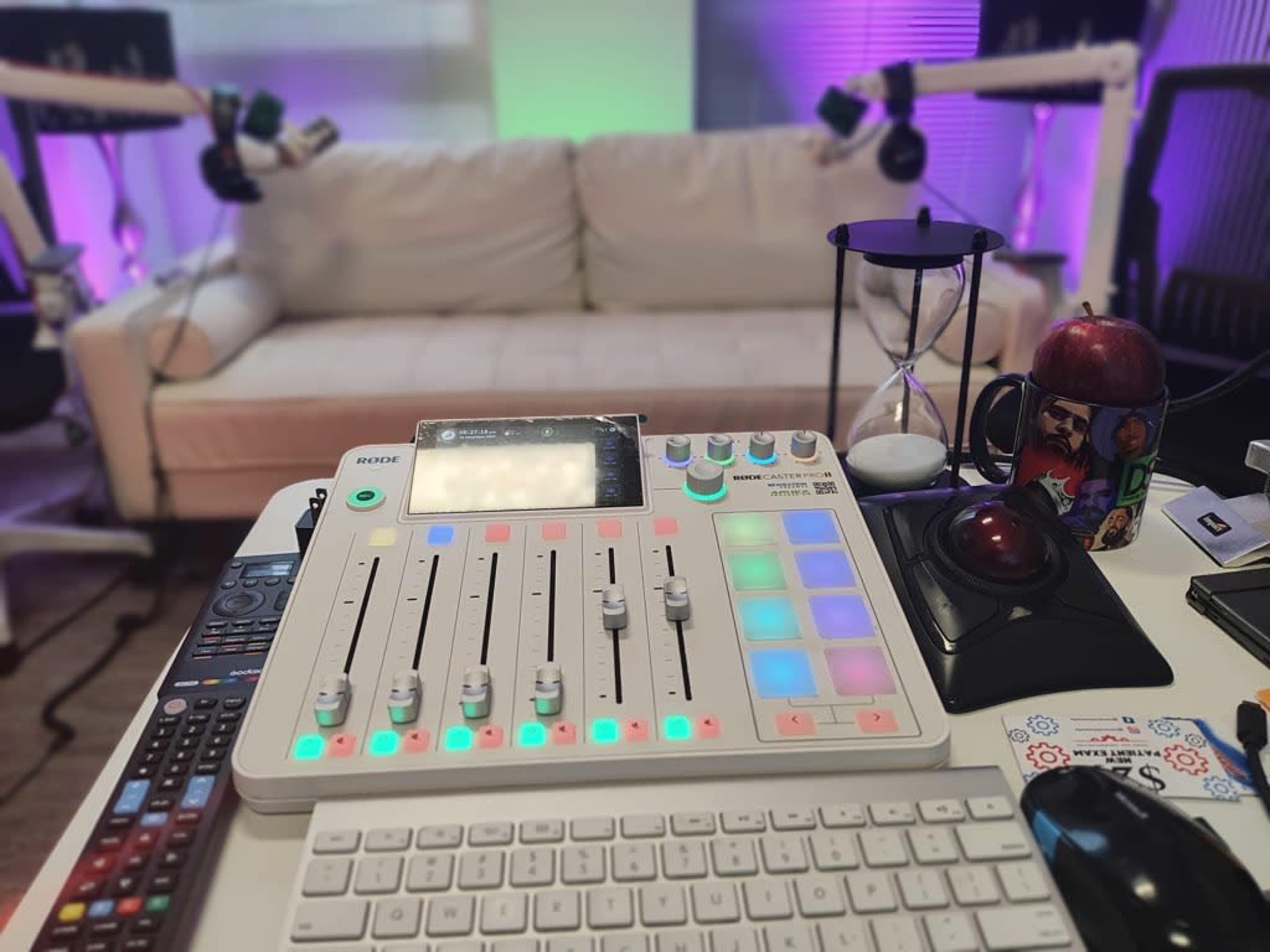 A digital audio mixer is positioned on a desk in front of a white couch, with colorful lighting in the background.
