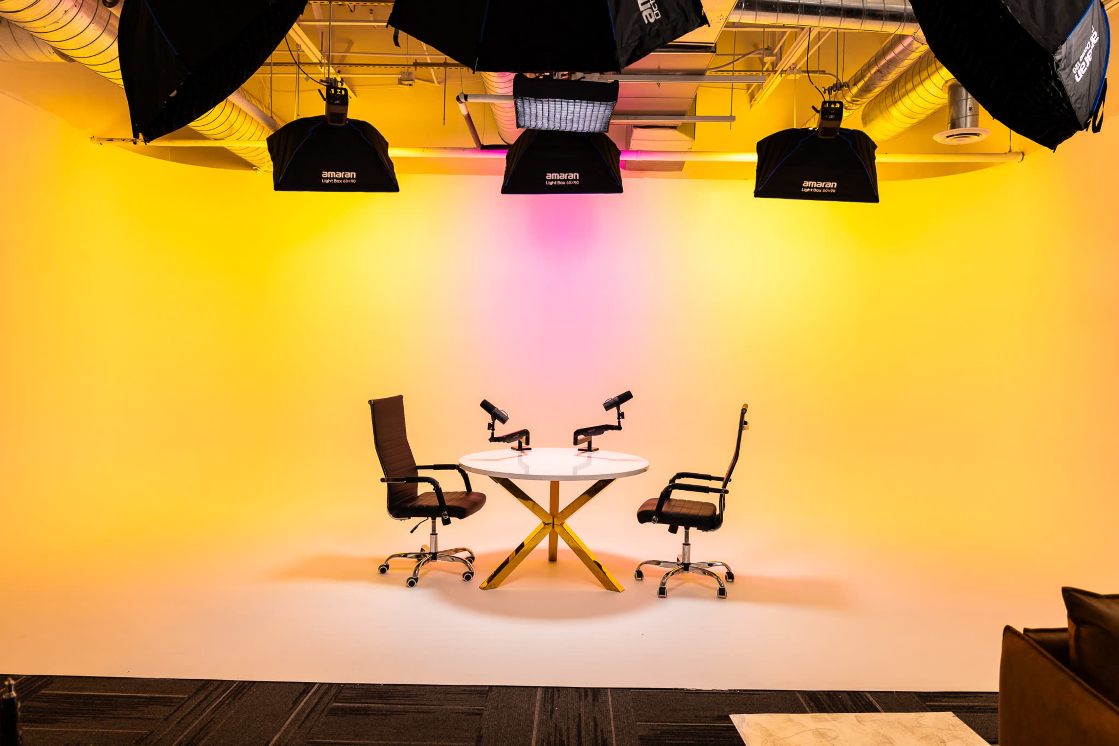 The image shows a modern studio setup with two chairs facing each other at a round table, under multiple studio lights against a vibrant gradient background.