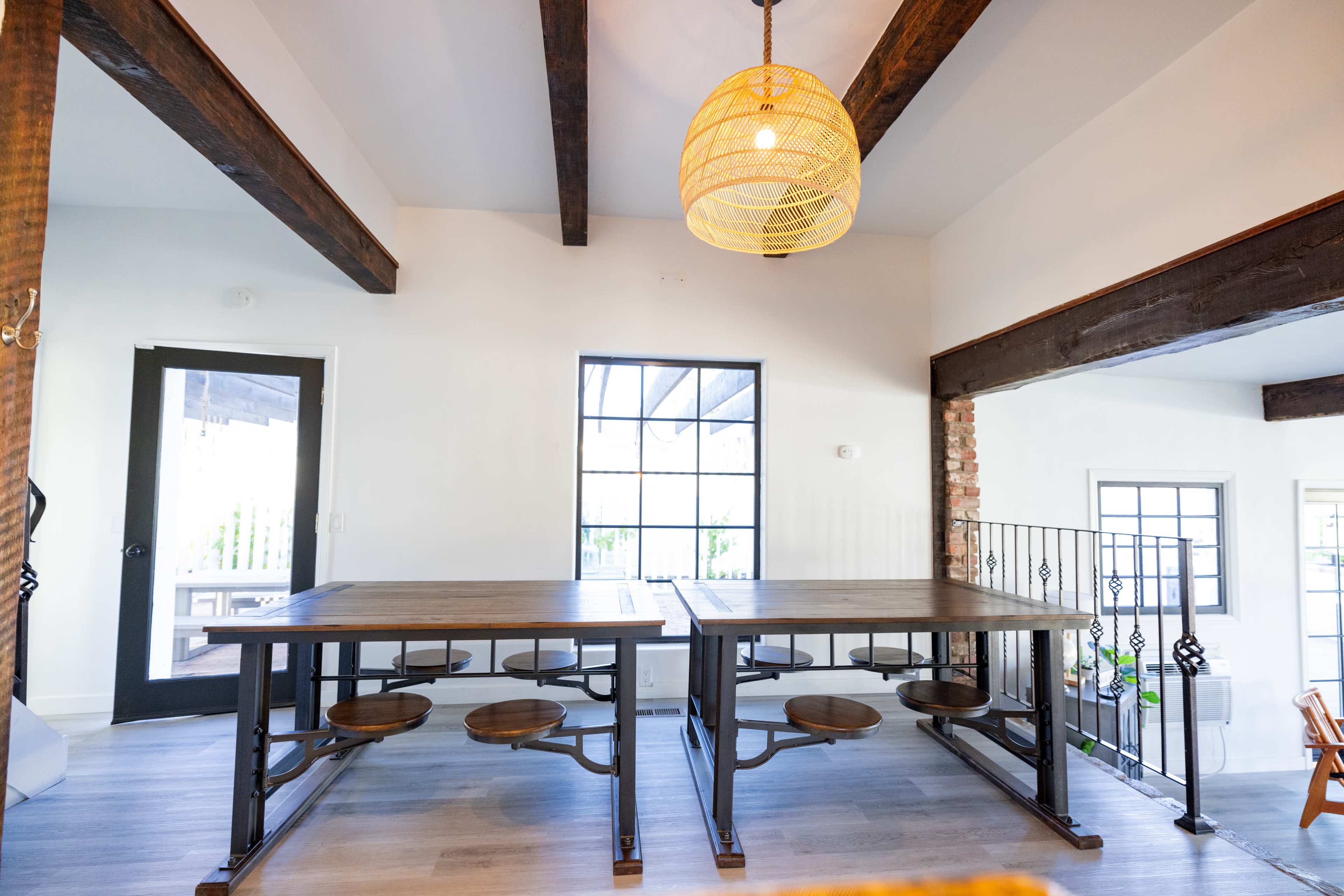 An open dining area features a large wooden table with circular wooden seats, illuminated by a woven pendant light, and surrounded by exposed wooden beams.