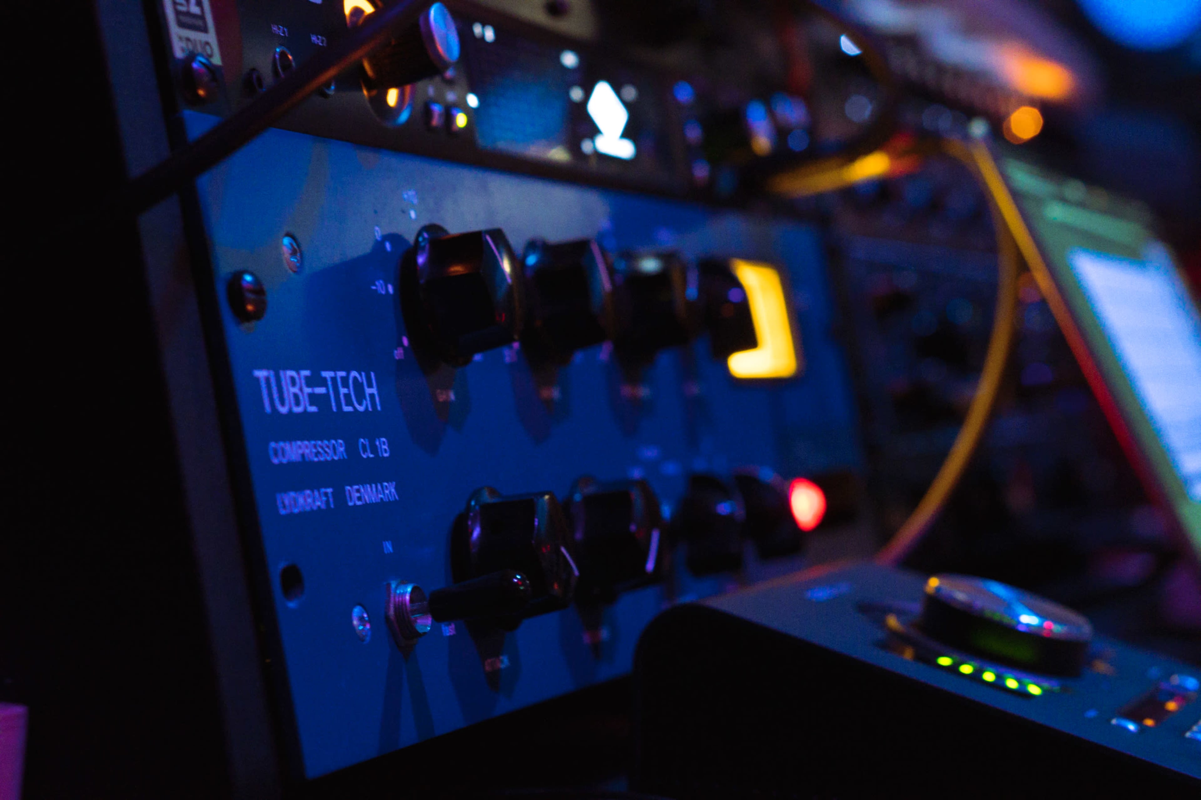The image shows a close-up of a blue audio compressor unit with labeled knobs and a digital display in a music production environment.