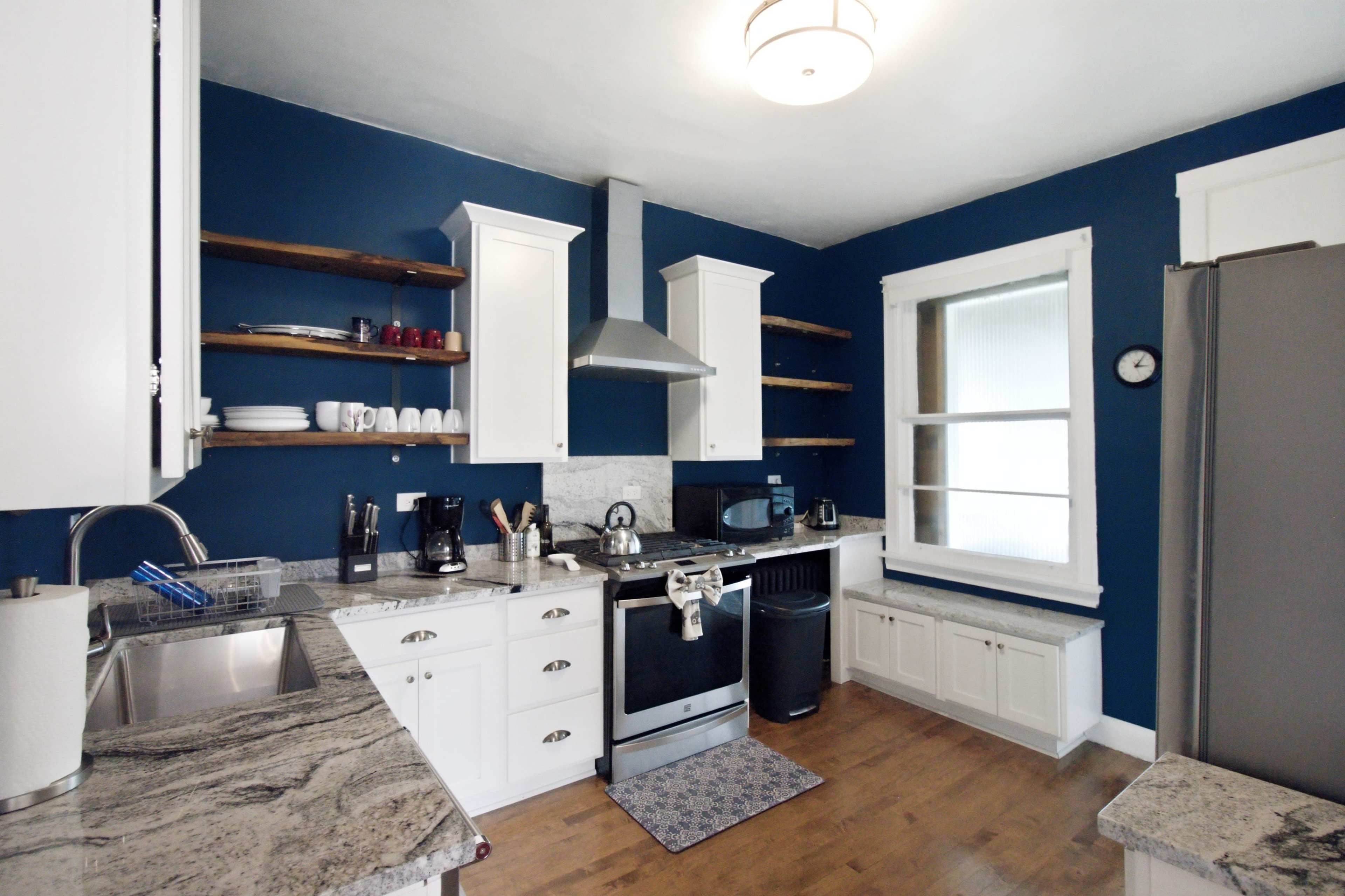 The image shows a modern kitchen with white cabinetry, open shelves, stainless steel appliances, and blue walls.